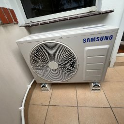 Serhii Cholavyn - Zamontowana jednostka zewnętrzna klimatyzacji Samsung na wspornikach, przy ścianie budynku z widoczną instalacją elektryczną w peszlu. Widok z dołu, blisko okna.