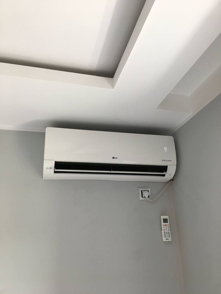 Biała klimatyzacja LG Dual Inverter zamontowana na szarej ścianie w pomieszczeniu z dekoracyjnym sufitem podwieszanym. Widoczny pilot i gniazdko elektryczne.