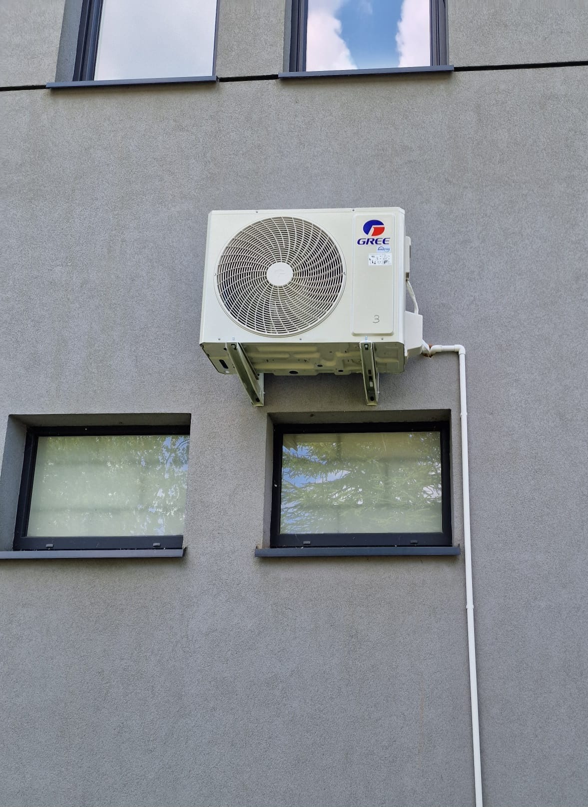Klimatyzator marki Gree zamontowany na szarej elewacji budynku, widoczny z boku, z rurą odprowadzającą. Dwa okna z czarnymi ramami po bokach i jedno u góry.