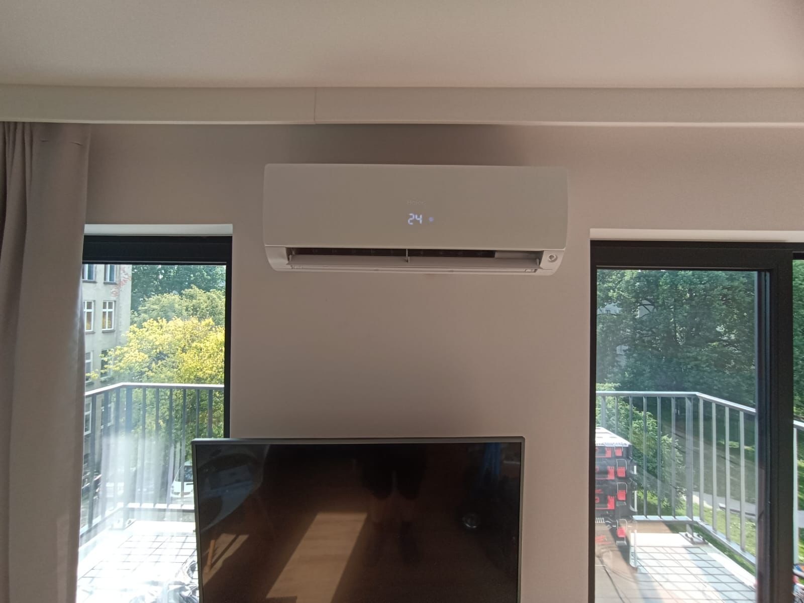 Biały klimatyzator ścienny z wyświetlaczem temperatury 24°C, zamontowany w jasnym pokoju z oknami balkonowymi i telewizorem. Widok na zieleń za oknem.