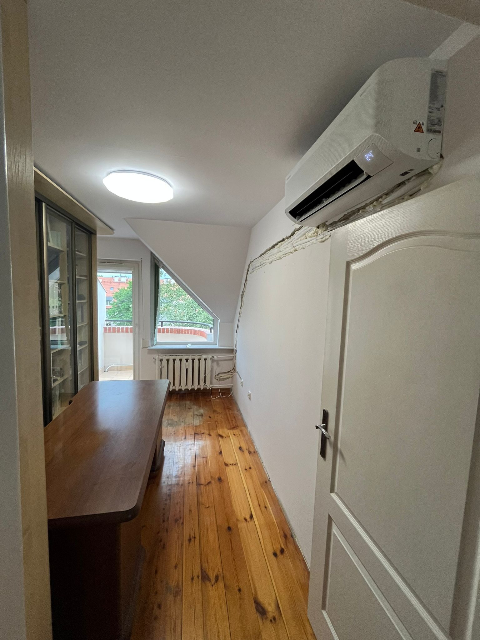 Biały klimatyzator na ścianie w pokoju na poddaszu, widoczna instalacja i uszczelnienie pianką montażową. Drewniana podłoga i okno balkonowe w tle.