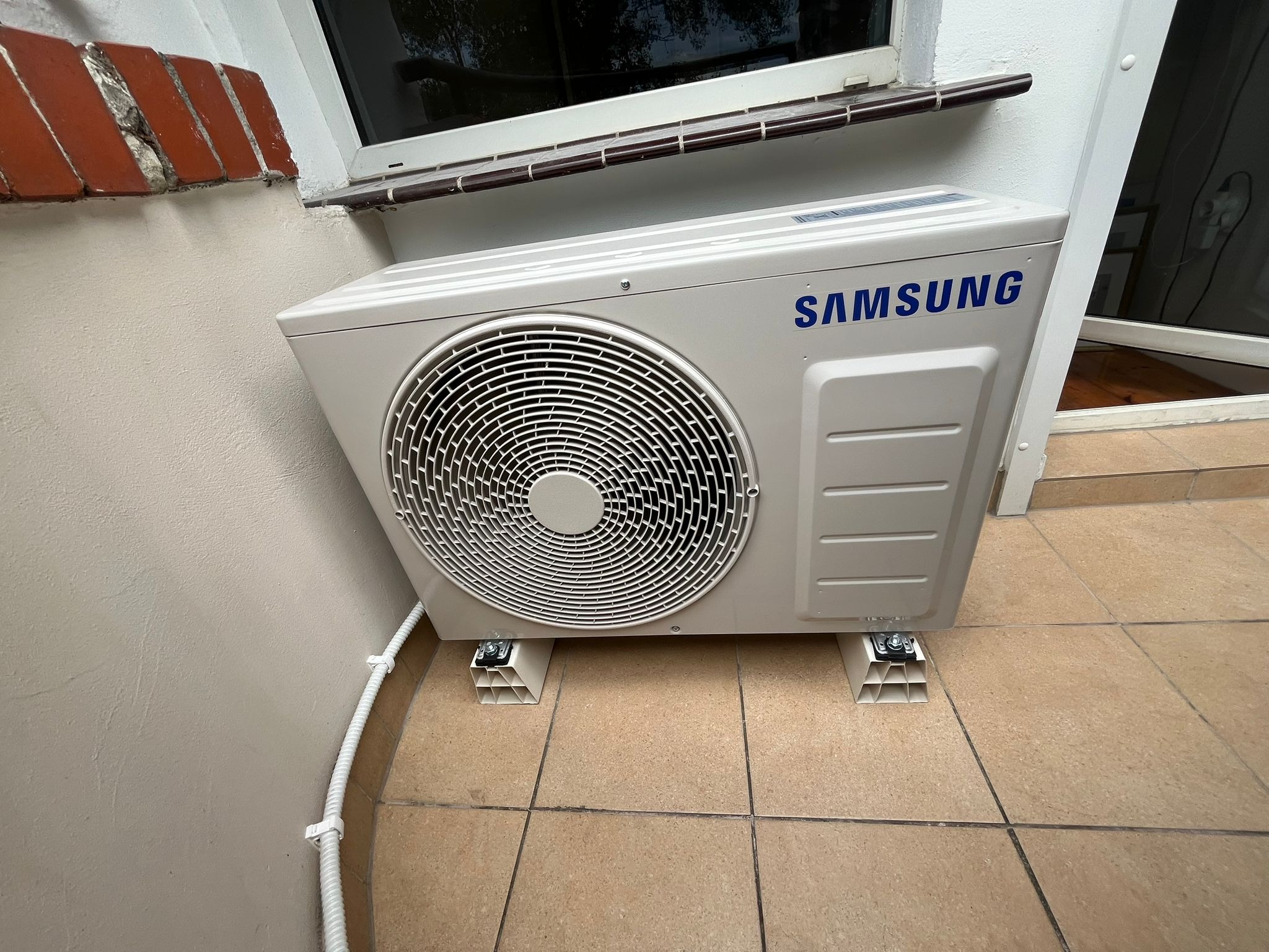 Zamontowana jednostka zewnętrzna klimatyzacji Samsung na wspornikach, przy ścianie budynku z widoczną instalacją elektryczną w peszlu. Widok z dołu, blisko okna.