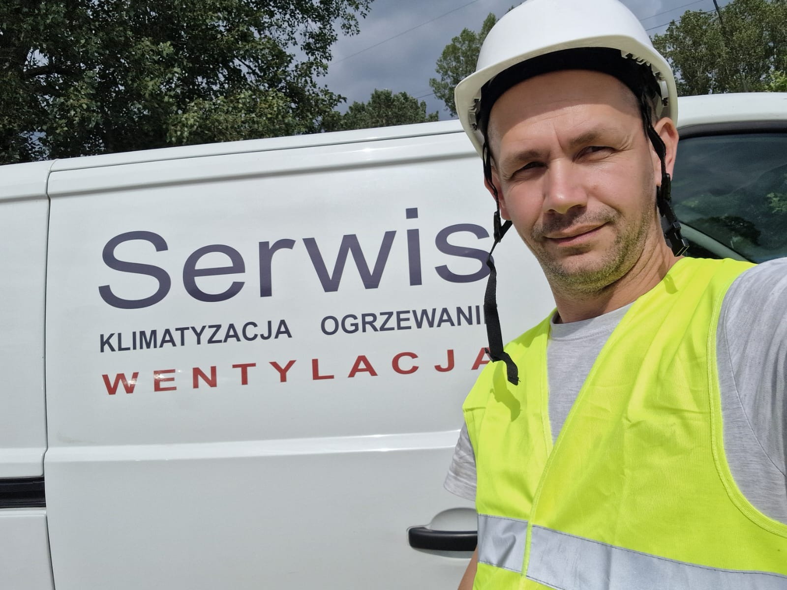 Pracownik serwisu klimatyzacji, ogrzewania i wentylacji w kamizelce odblaskowej i kasku na tle firmowego samochodu w Warszawie. Widoczne logo firmy.