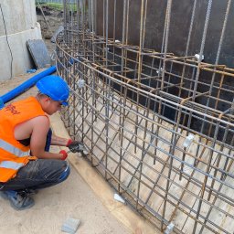 Oleksii Semeniuk - Pracownik w kasku i kamizelce montuje zbrojenie z prętów stalowych, tworząc szkielet konstrukcji na fundamencie. Widoczne narzędzia i zabezpieczenia.