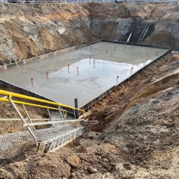 Oleksii Semeniuk - Zalany fundament pod budowę z wystającymi elementami zbrojenia, widok z góry z metalowymi schodami na pierwszym planie i częściowo widoczną betoniarką w tle.