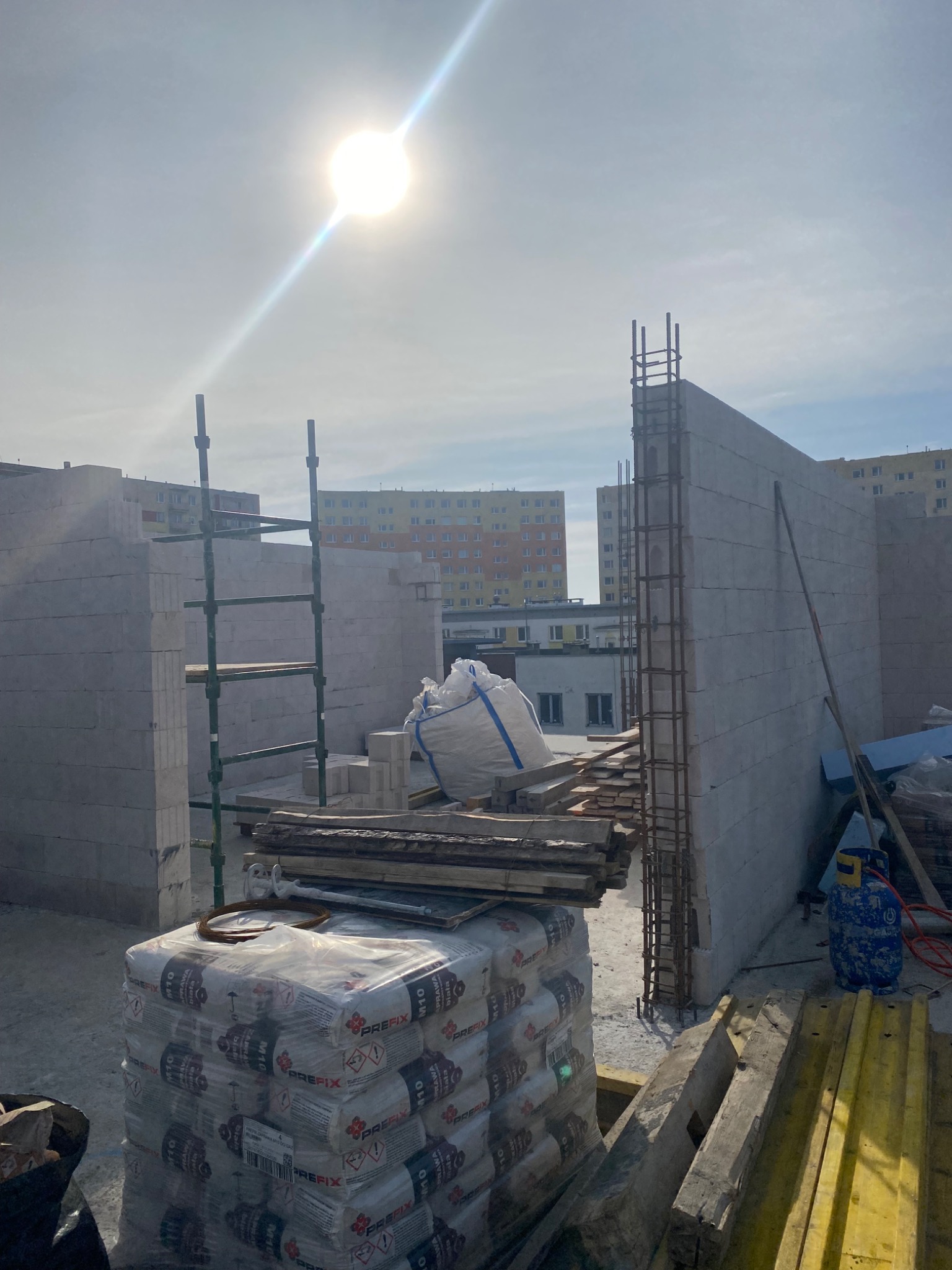 Budowa ścian z betonu komórkowego w słoneczny dzień. Widoczne zbrojenia, palety z zaprawą murarską, rusztowanie i otoczenie budowy z blokami.