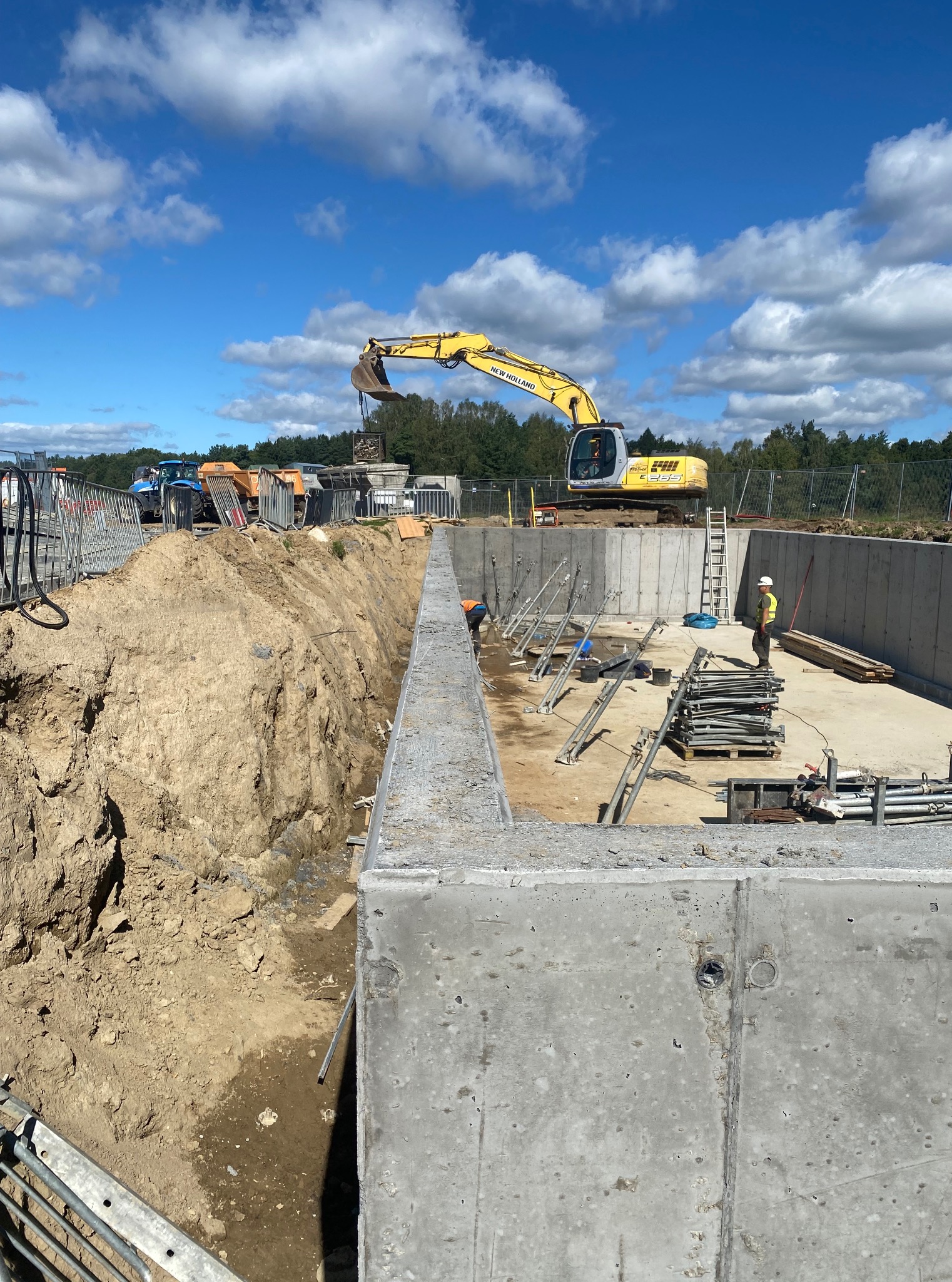 Wykop pod fundamenty z betonowymi ścianami szalunkowymi, na budowie stoi koparka New Holland i pracownik w kasku. Błękitne niebo z chmurami w tle.