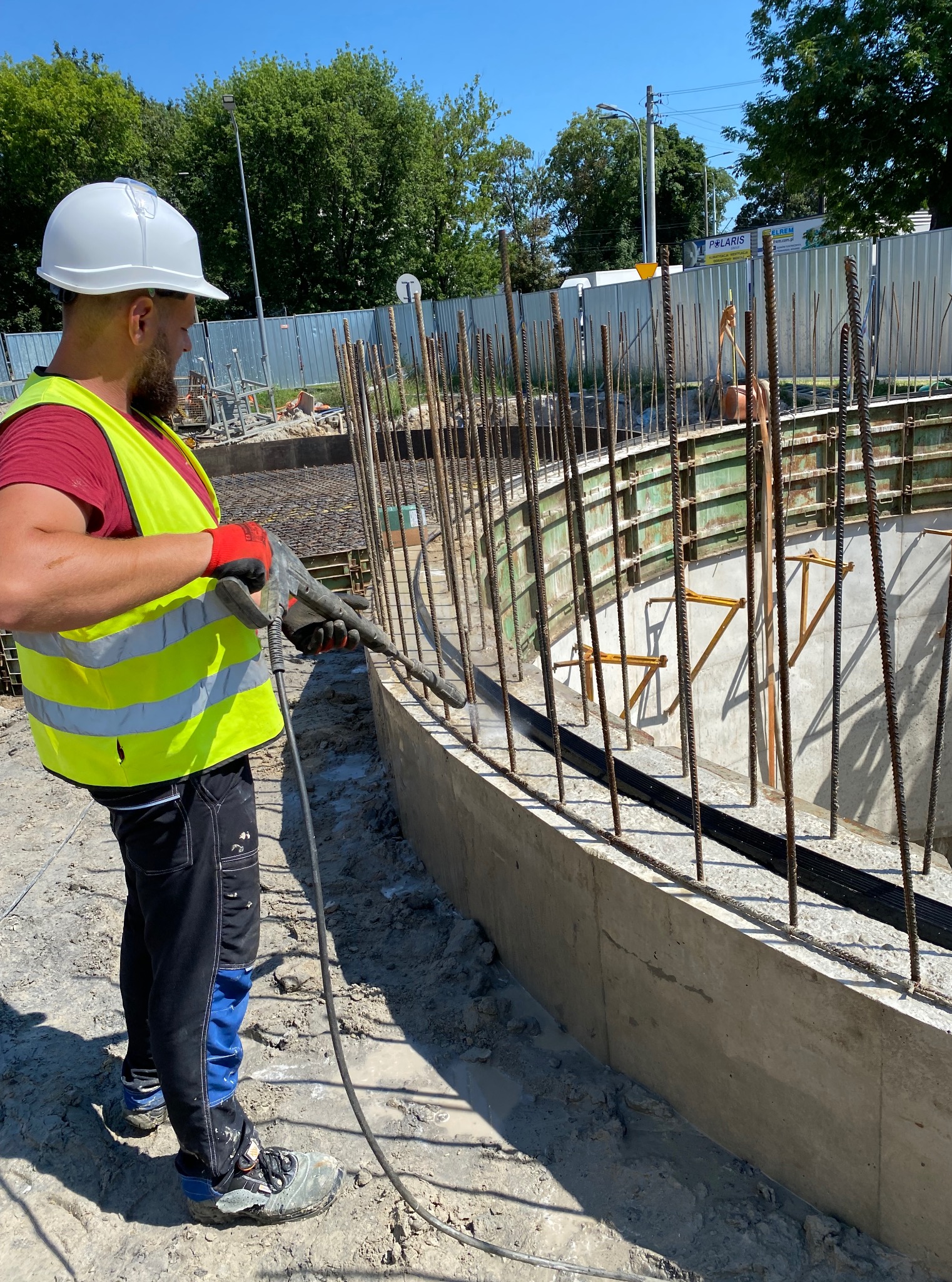 Pracownik w kasku i kamizelce, z pistoletem do betonu, pracuje przy zbrojeniu fundamentów. Widoczne szalunki i stalowe pręty zbrojeniowe. Słoneczny dzień na budowie.