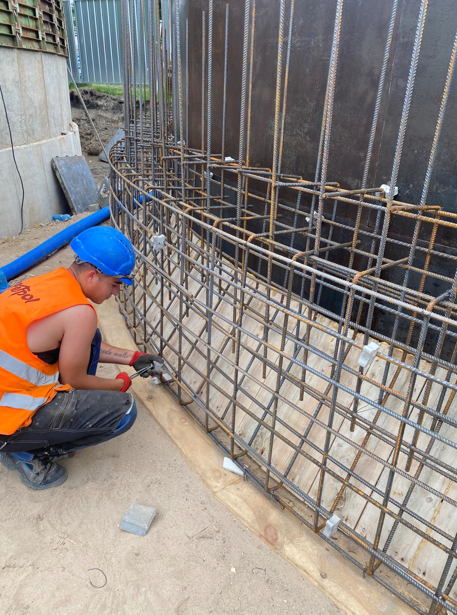 Pracownik w kasku i kamizelce montuje zbrojenie z prętów stalowych, tworząc szkielet konstrukcji na fundamencie. Widoczne narzędzia i zabezpieczenia.