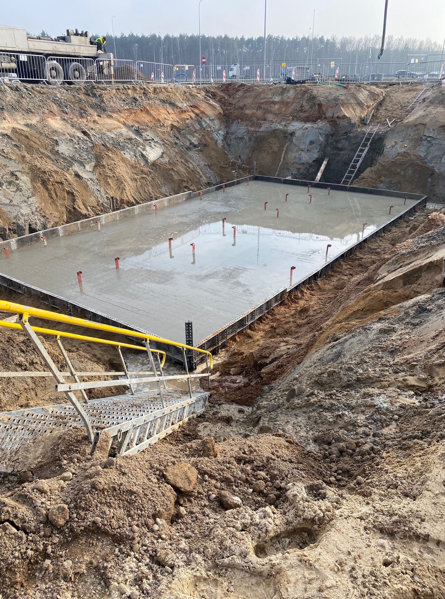 Zalany fundament pod budowę z wystającymi elementami zbrojenia, widok z góry z metalowymi schodami na pierwszym planie i częściowo widoczną betoniarką w tle.