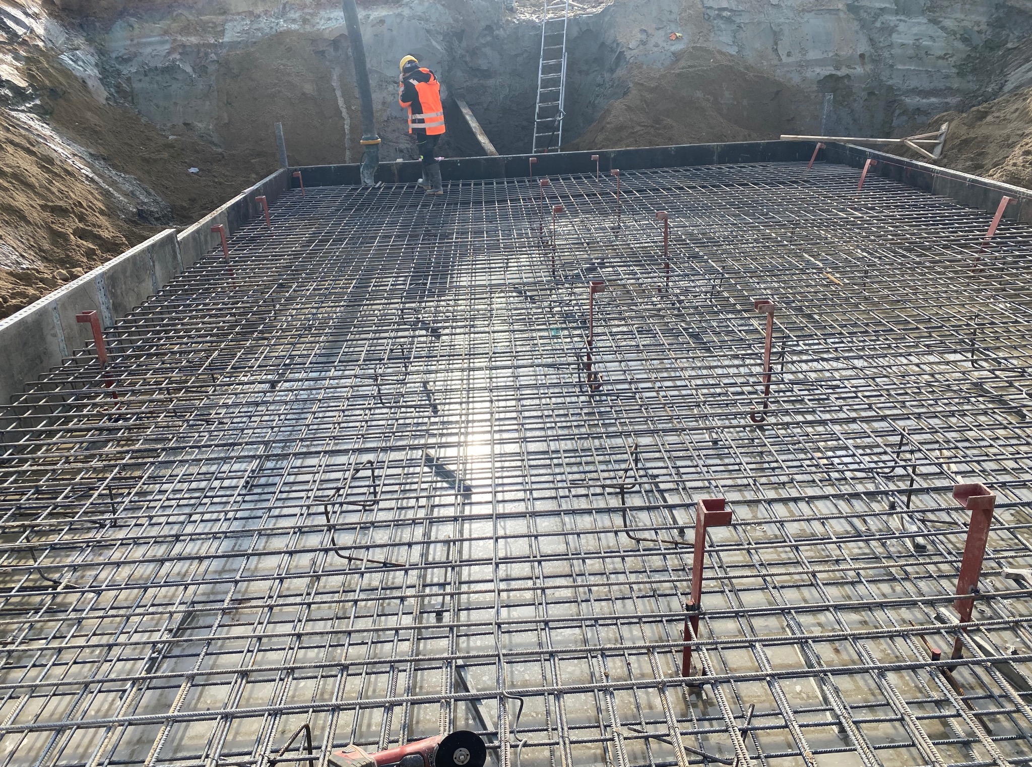 Zbrojenie fundamentu płyty betonowej z widocznymi prętami, podkładkami dystansowymi i pracownikiem w odblaskowej kamizelce na tle wykopu i drabiny.