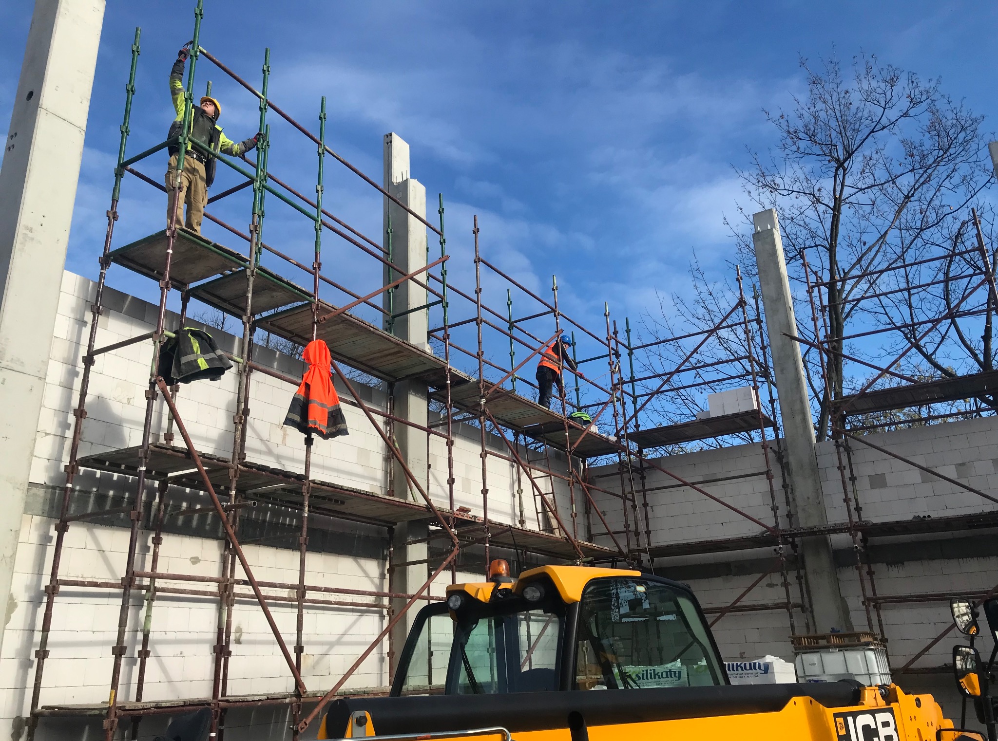 Budowa domu z białych bloczków silikatowych, widoczni dwaj pracownicy na rusztowaniu. Na pierwszym planie żółta maszyna budowlana.