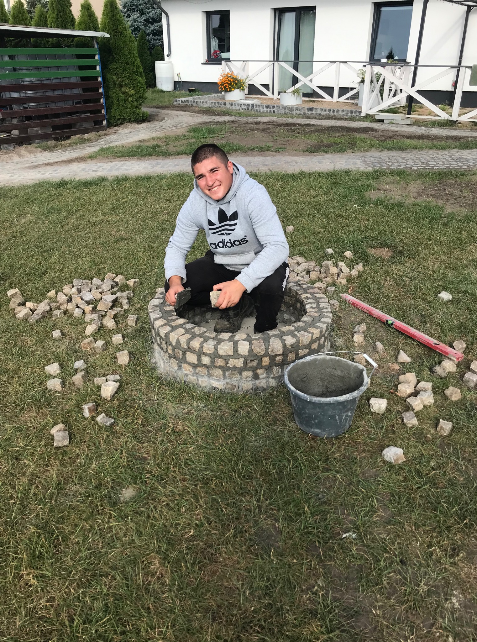 Młody mężczyzna w bluzie Adidas muruje kamienny krąg na trawniku, obok leżą narzędzia i wiadro z zaprawą. W tle biały dom z płotem.