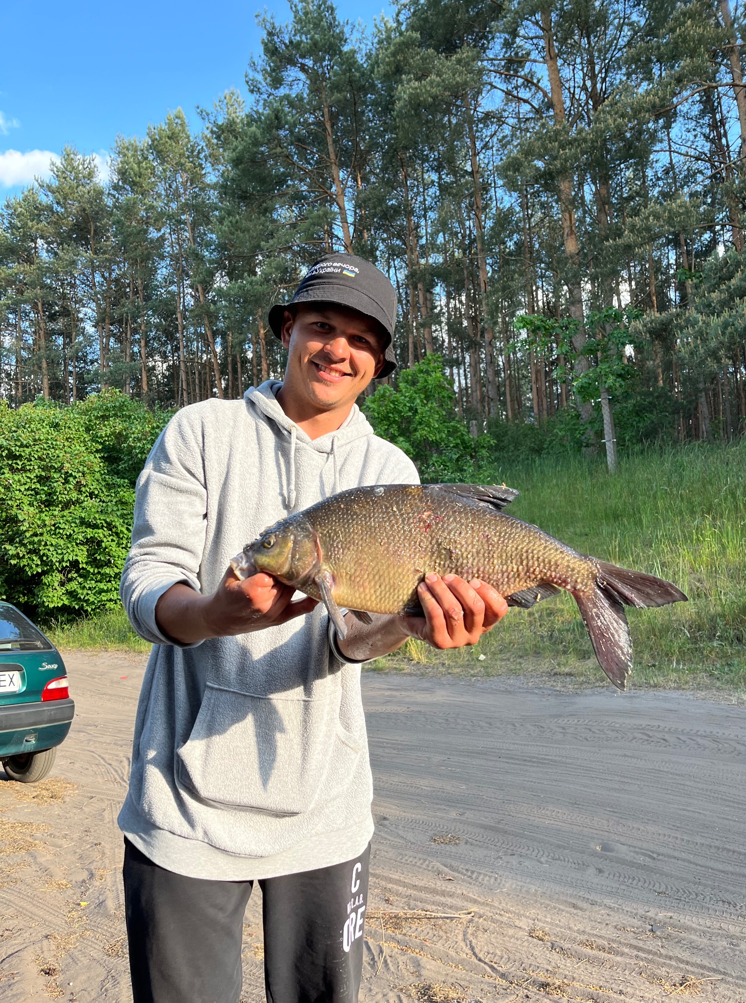 Uśmiechnięty mężczyzna w czarnym kapeluszu trzyma dużego karpia na tle lasu i zielonego samochodu. Słoneczny dzień.