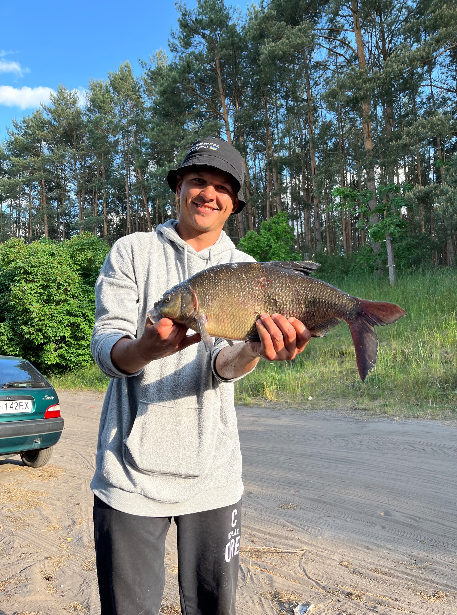 Uśmiechnięty mężczyzna w kapeluszu prezentuje złowioną rybę na tle leśnym i zielonego auta. Połyskujące łuski karpia w promieniach słońca.