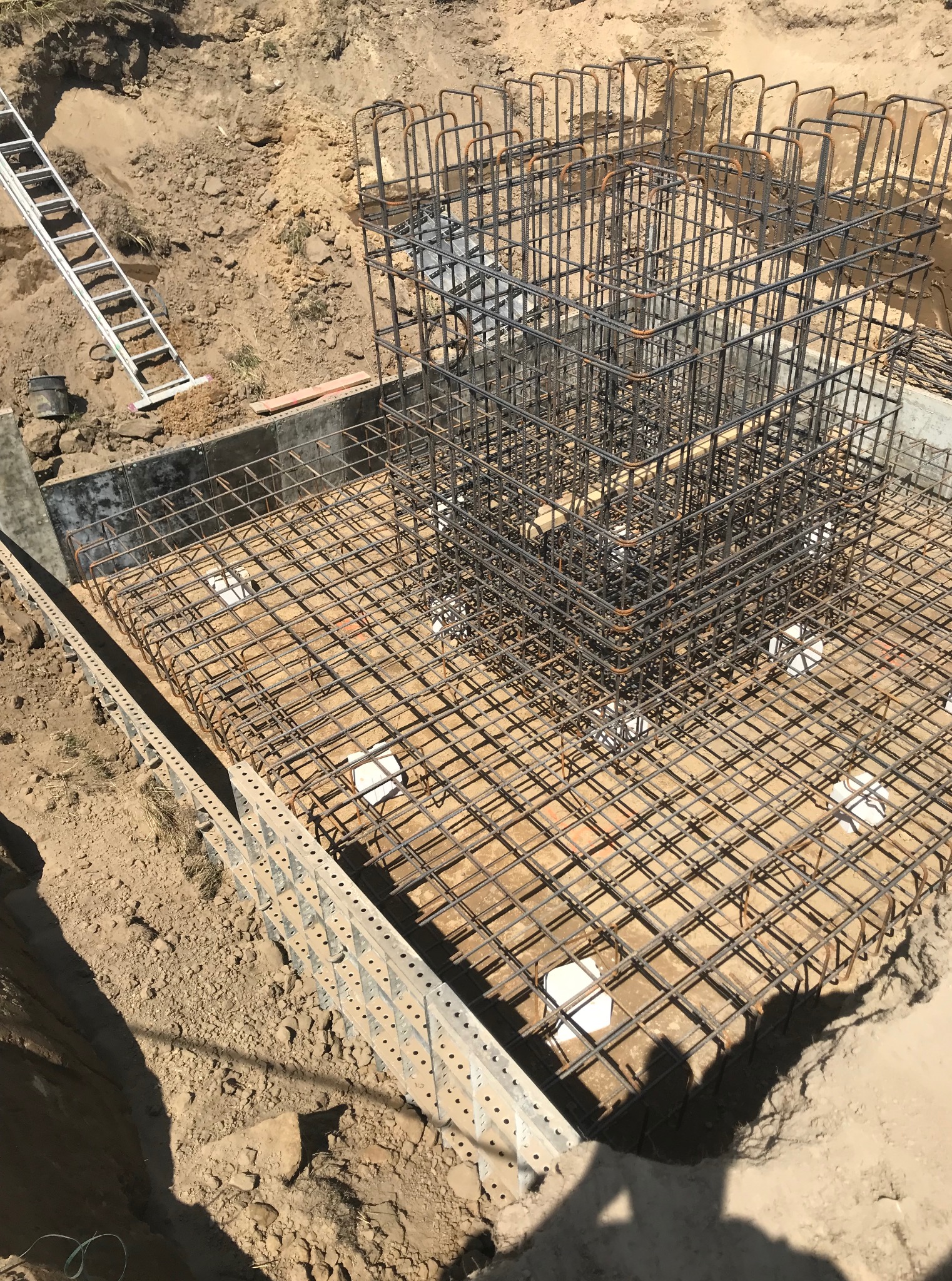 Zbrojenie fundamentu pod budynek, widoczne stalowe pręty tworzące szkielet, białe podkładki dystansowe i szalunek. Na ziemi leży drabina.