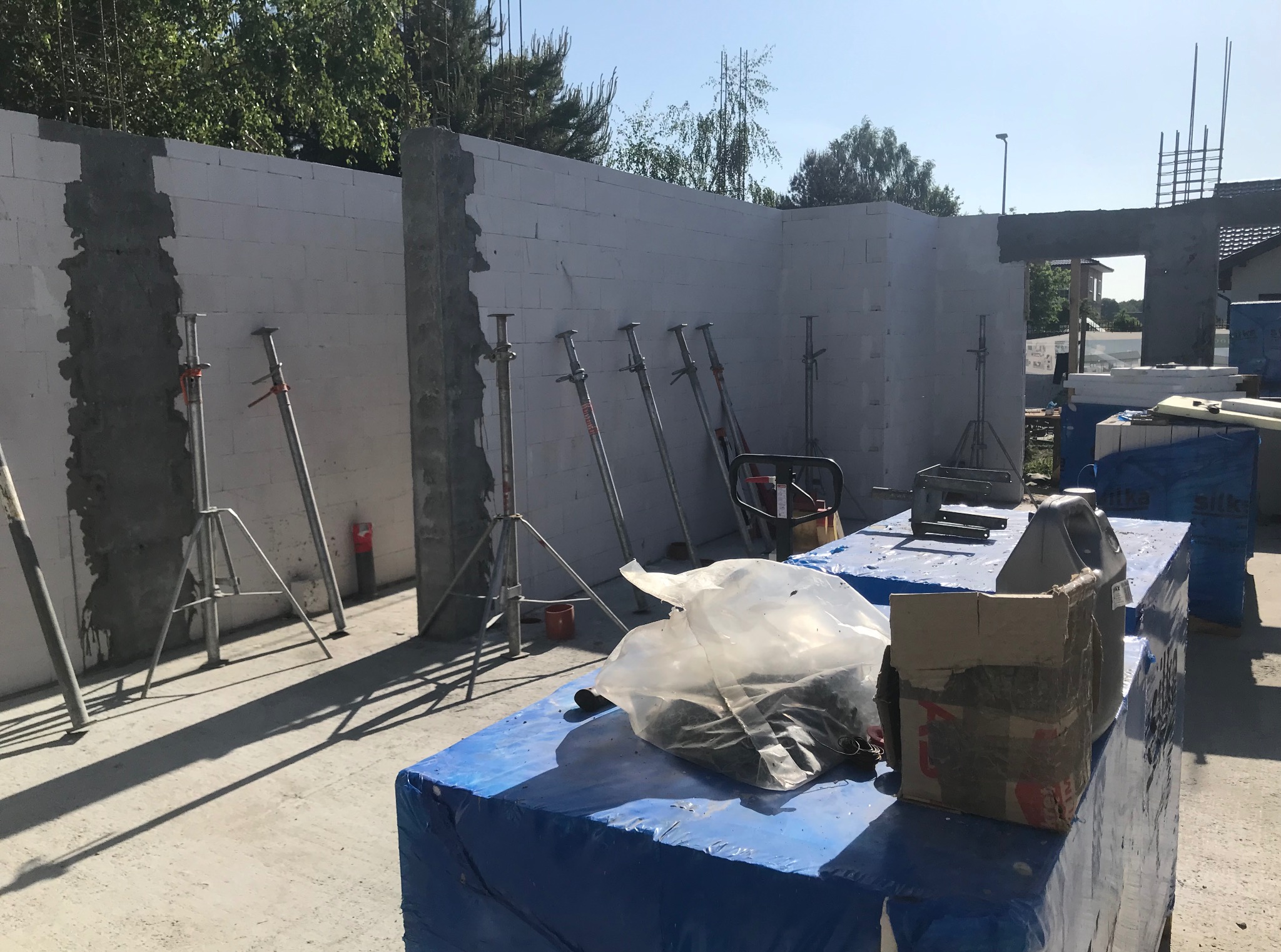 Wznoszone ściany z białych bloczków, wsparte metalowymi stemplami. Widoczne materiały budowlane, fragment konstrukcji zbrojeniowej i zarys budynku w tle.