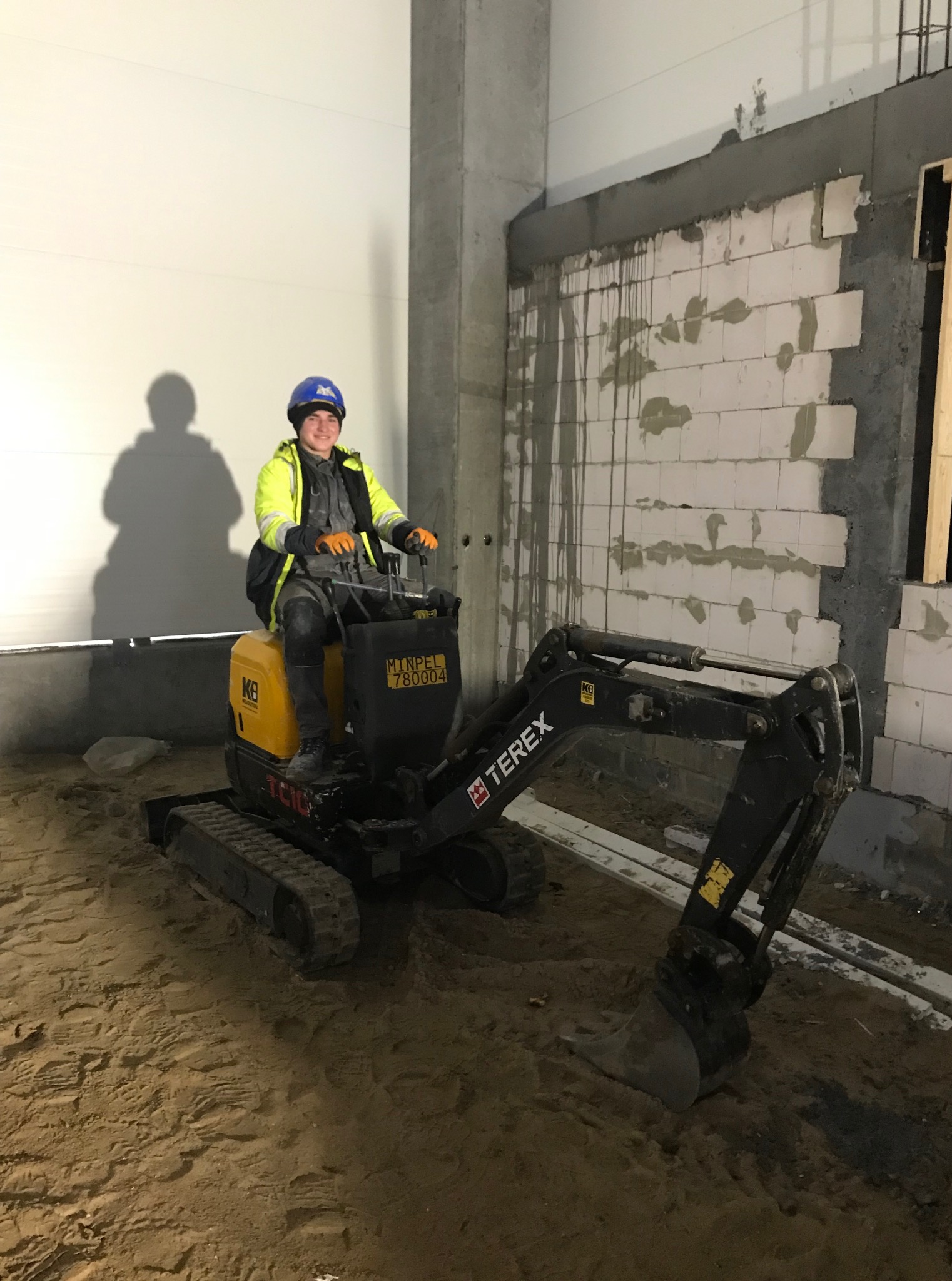 Operator w kasku na mini koparce Terex, na tle murowanej ściany z betonu komórkowego. W tle cień osoby na ścianie.