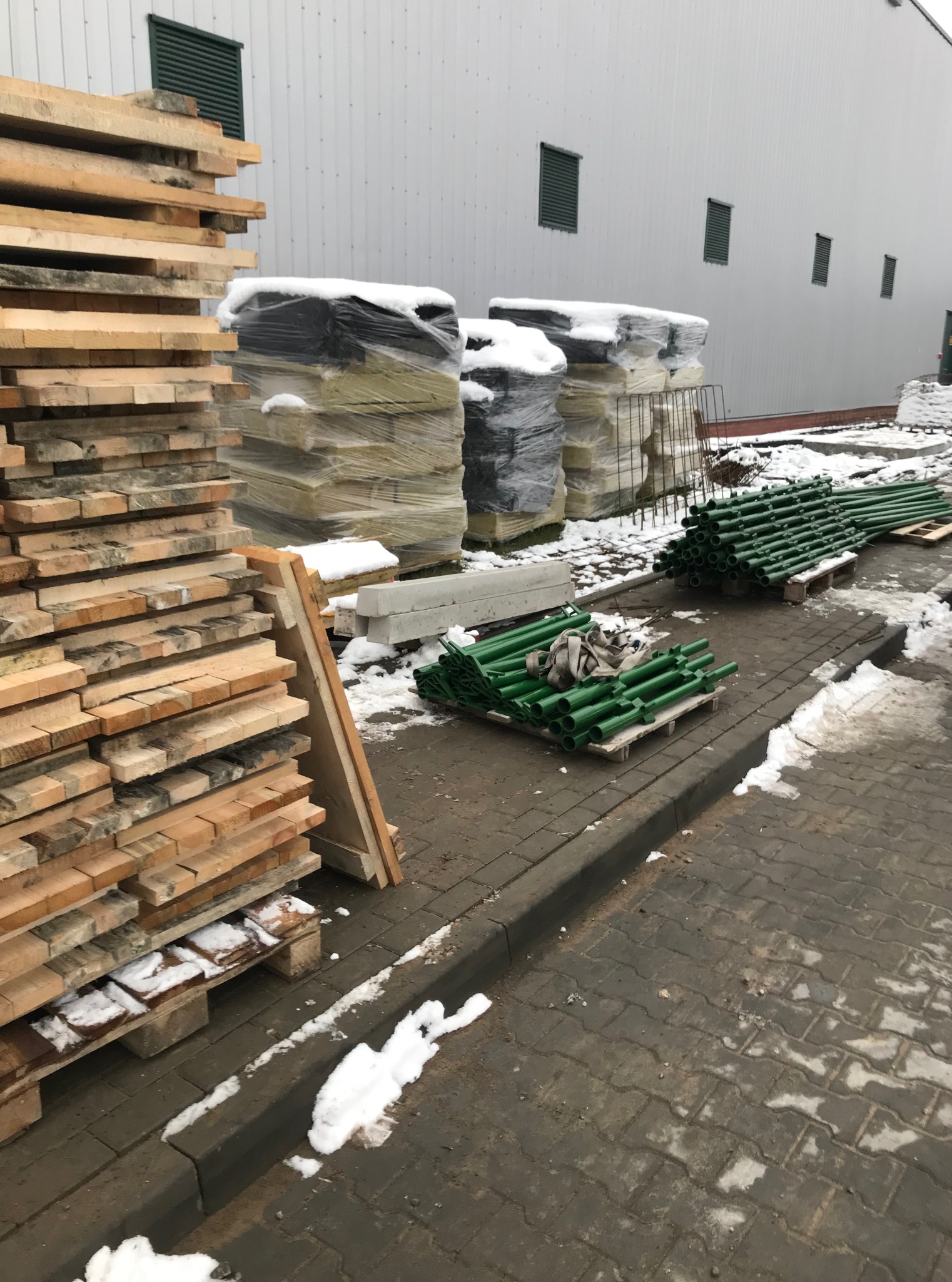 Składowane materiały budowlane na zewnątrz: drewno, ocieplenie w folii, betonowe elementy i zielone podpory budowlane, częściowo pokryte śniegiem.
