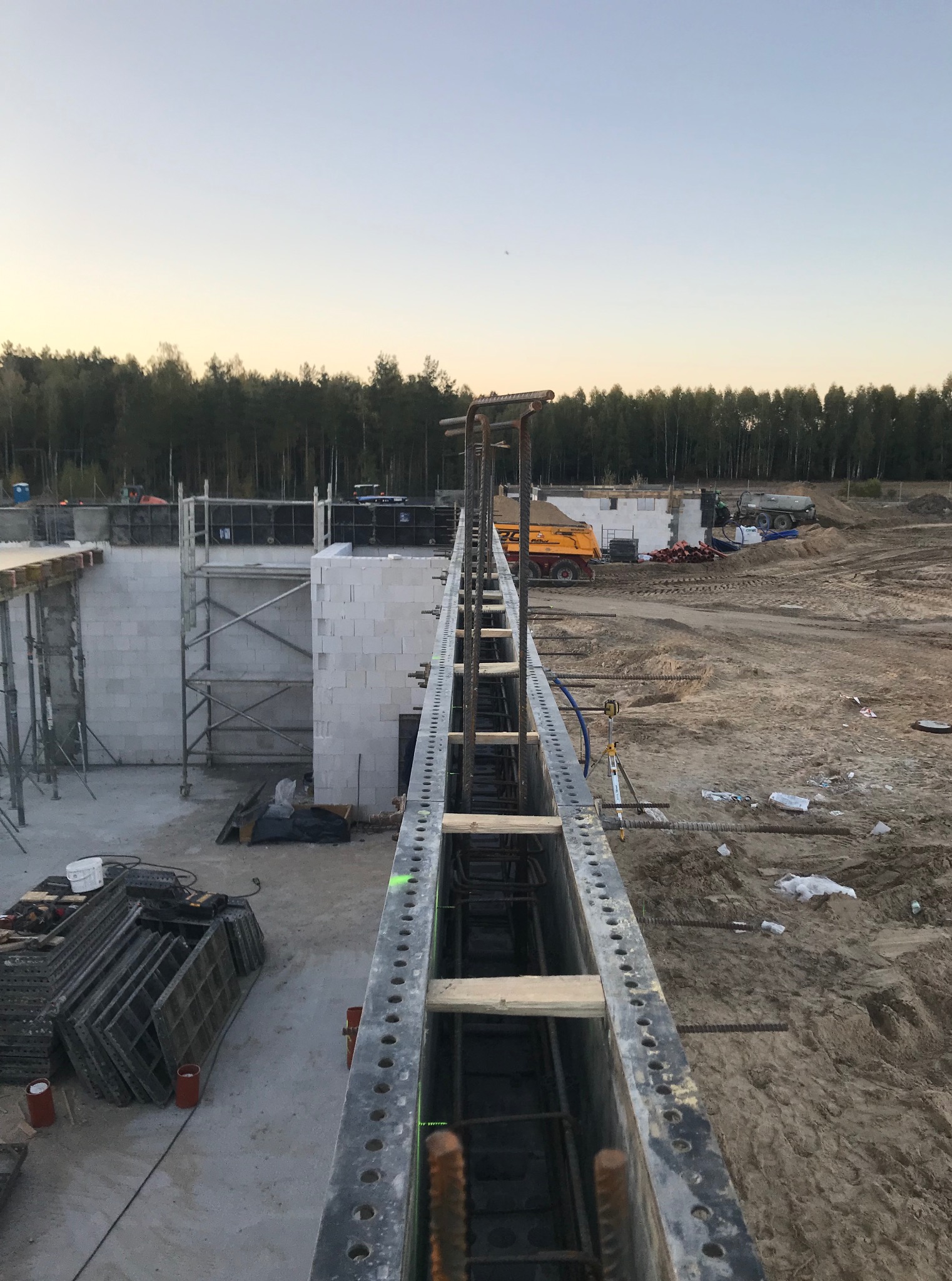 Budowa fundamentów: szalunek zbrojony stalą na tle wzniesionych ścian z bloczków, sprzęt budowlany i las w oddali, ujęcie z góry, wczesny etap prac.