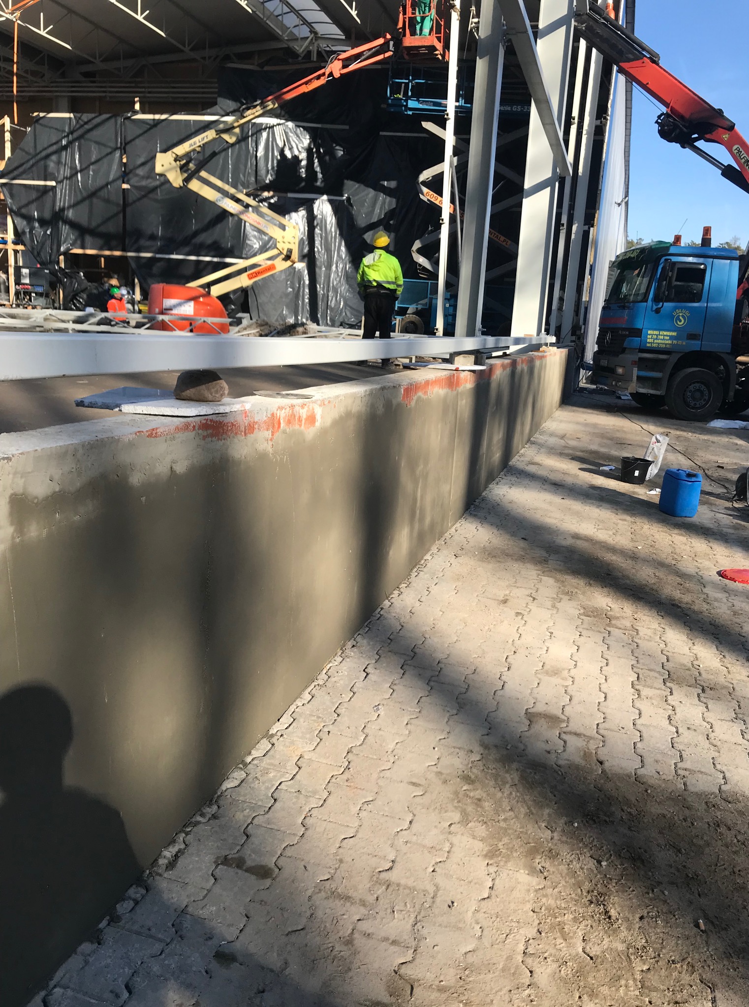 Fundament budynku przemysłowego po wylaniu, z pracownikiem w żółtej kamizelce i sprzętem budowlanym w tle. Widoczny fragment ciężarówki z dźwigiem.