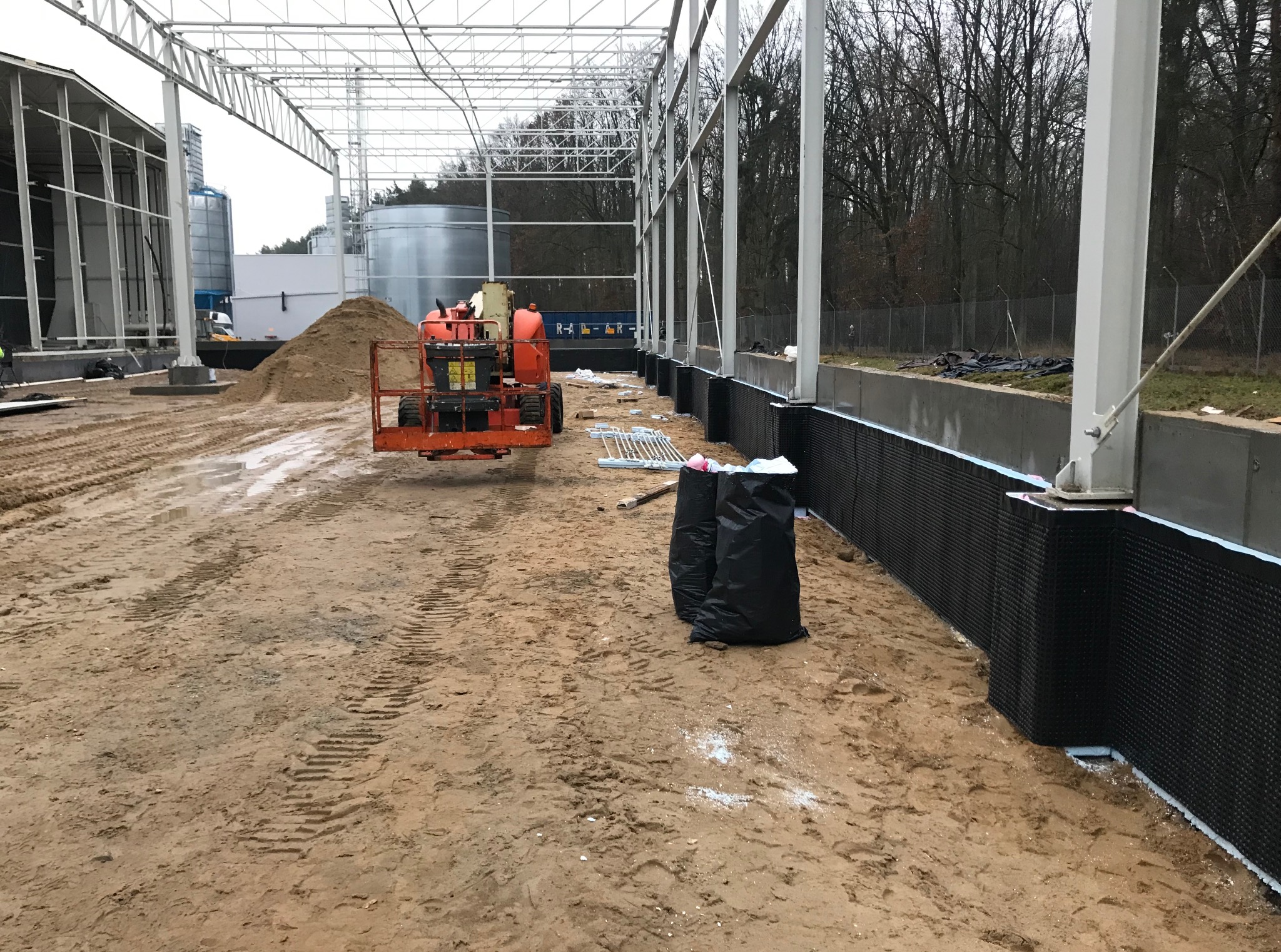 Budowa hali: widok na fundamenty z izolacją, stalową konstrukcję i podnośnik. Na ziemi ślady opon i hałda piasku. W tle drzewa.