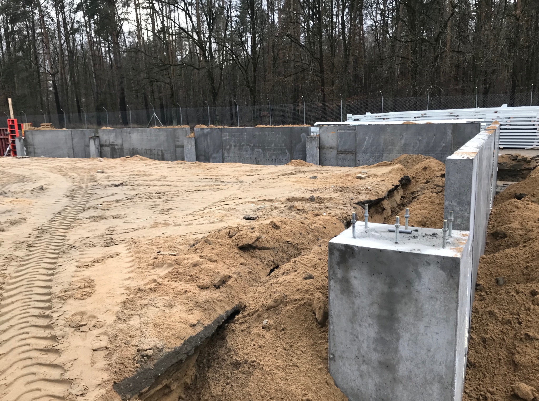 Wykop pod fundamenty z widocznymi betonowymi ścianami, wystającymi śrubami i śladami opon na piasku. W tle las i elementy szalunków.