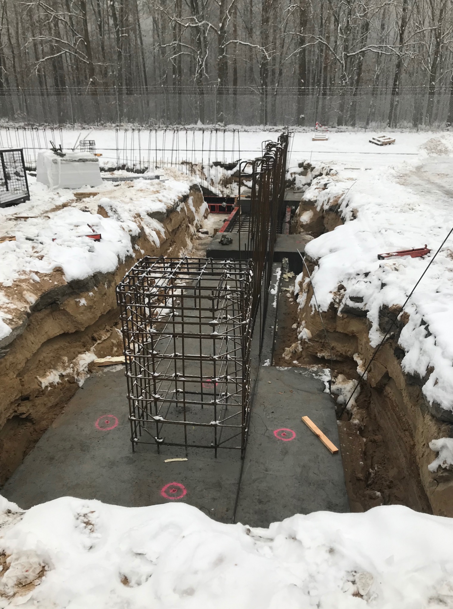 Zimowy wykop pod fundamenty z widocznym zbrojeniem i betonową wylewką. Śnieg leży na krawędzi wykopu i w tle. Zbrojenie w kształcie klatki na pierwszym planie.