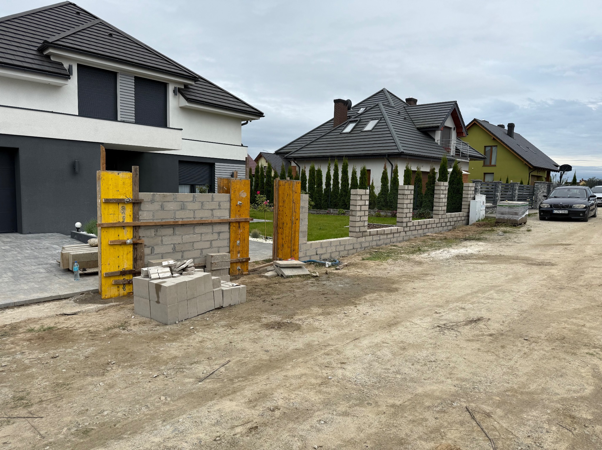 Wznoszenie muru z bloczków betonowych na posesji z nowymi domami. Żółte szalunki, widoczne bloczki i fragmenty muru. Prace murarskie w toku, krajobraz podmiejski.