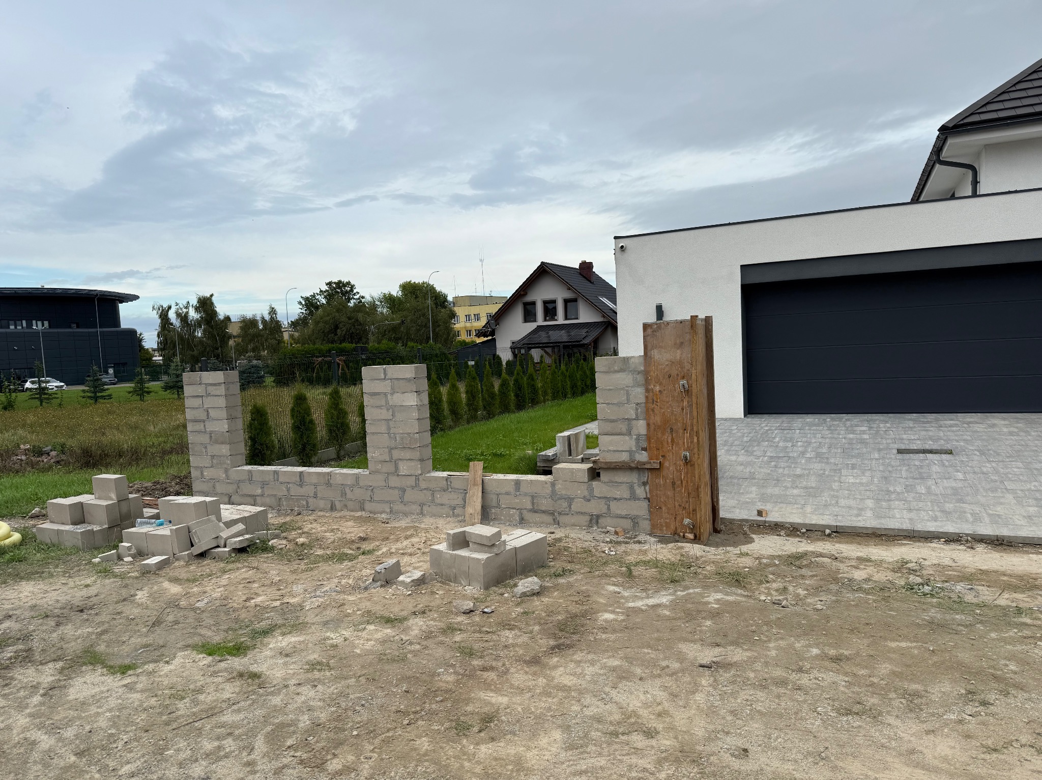Wznoszenie ściany z bloczków betonowych na tle nowoczesnego domu z garażem. Widoczne niedokończone fragmenty muru i materiały budowlane na placu budowy.
