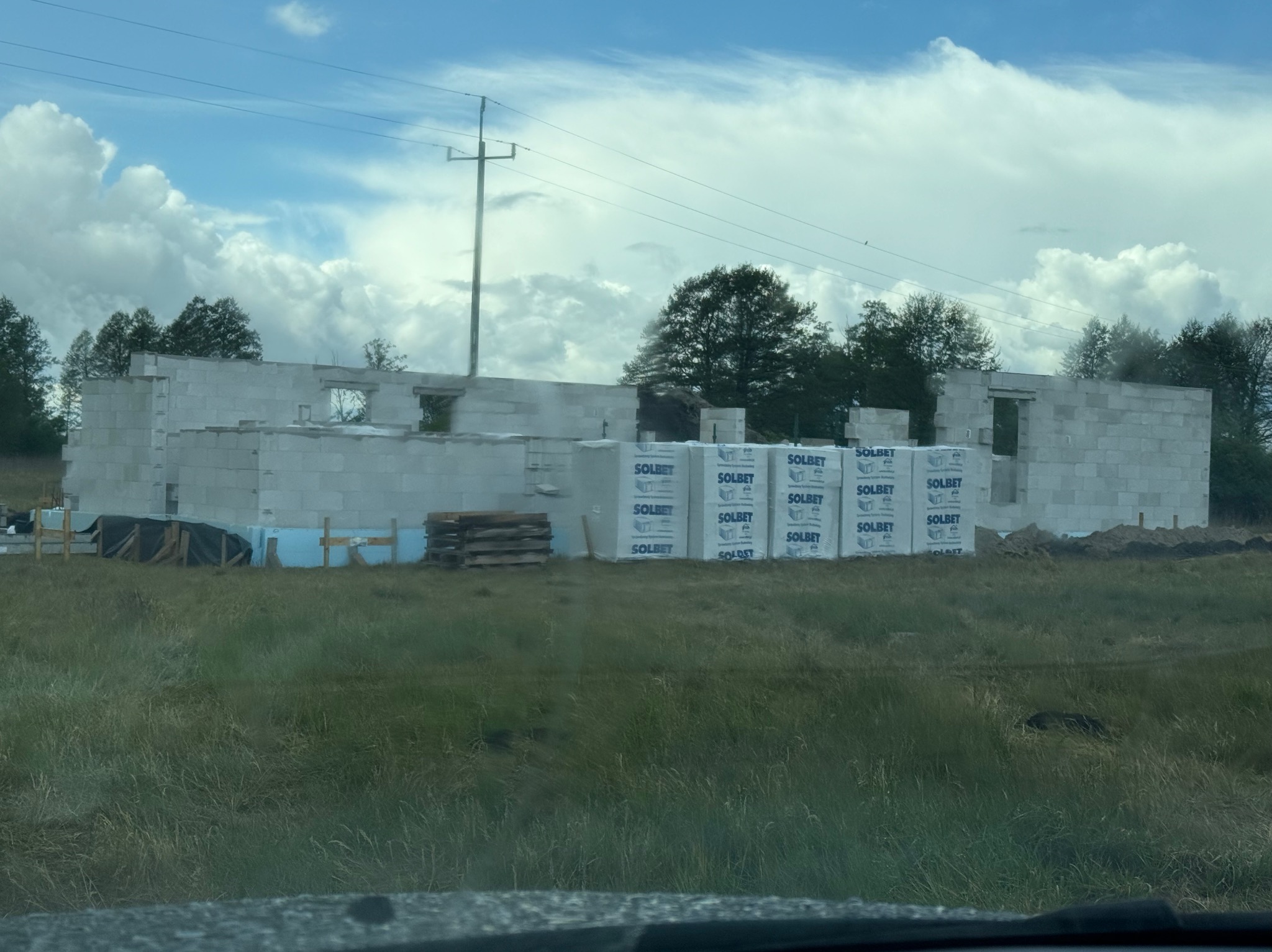 Wznoszone ściany domu z betonu komórkowego Solbet, widoczne palety z bloczkami. Budowa w otoczeniu zieleni, pod błękitnym niebem z obłokami.