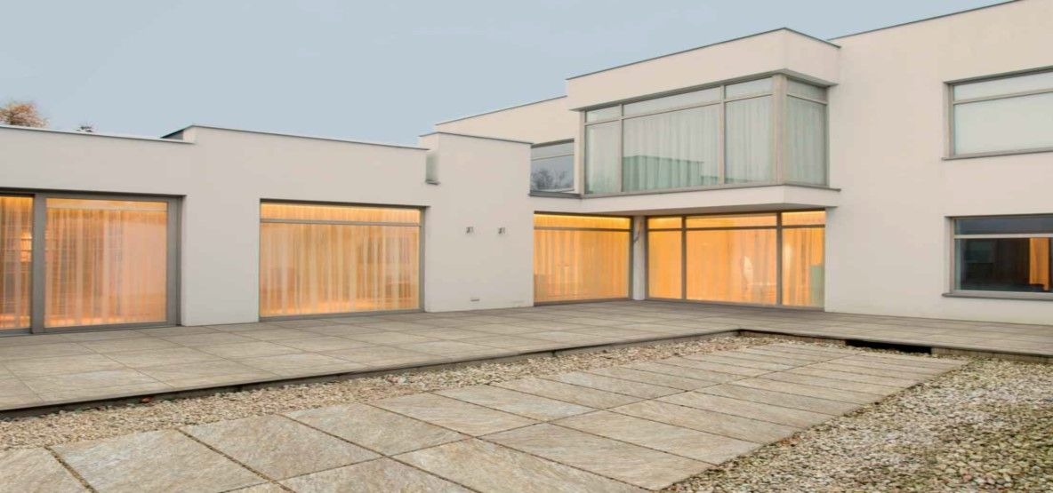 Nowoczesny dom z dużymi oknami i tarasem wyłożonym płytami w odcieniach beżu. Widoczne zasłony w oknach i żwirowe rabaty.