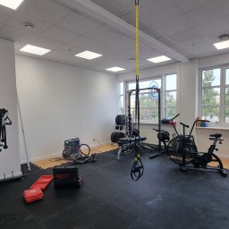 Oleksandr Mykharskyi - Jasne wnętrze siłowni z różnorodnym sprzętem do ćwiczeń: maszyna wyciągowa, rowerek, TRX, ciężary. Widoczne narzędzia sugerują trwające prace wykończeniowe.