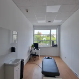 Jasne wnętrze gabinetu z kozetką, biurkiem przy oknie i umywalką. Parkiet na podłodze, sufit kasetonowy. Widok na zieleń za oknem.