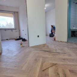 WektorinIN - Ułożona podłoga z paneli w jodełkę w remontowanym mieszkaniu. Widoczne narzędzia, klej i fragment niezakończonej podłogi. W tle wykończona łazienka.