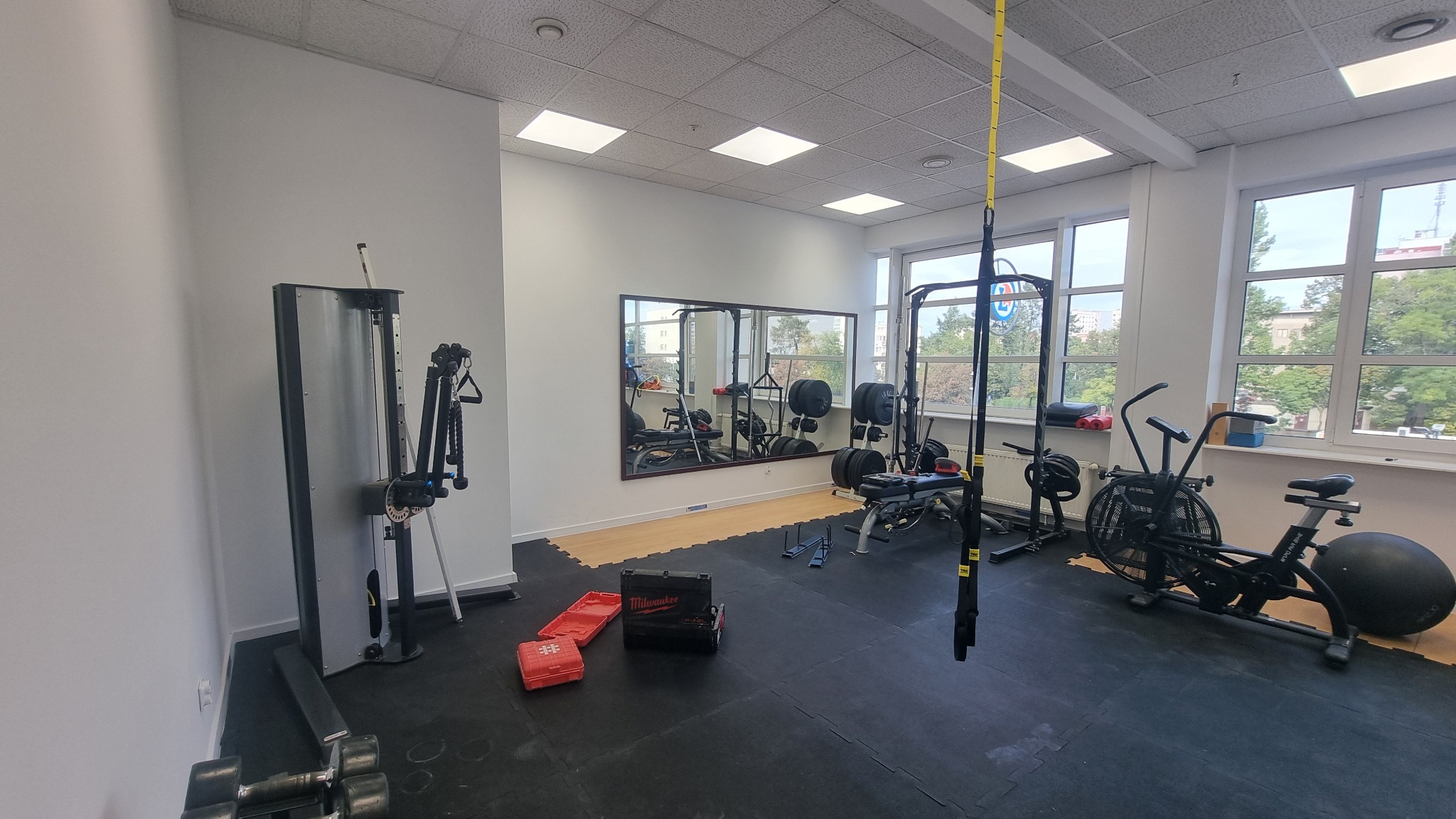 Jasne wnętrze siłowni z różnorodnym sprzętem: maszyna do ćwiczeń, rower stacjonarny, lustro, obciążenia i skrzynka narzędziowa. Widok na zewnątrz przez okna.