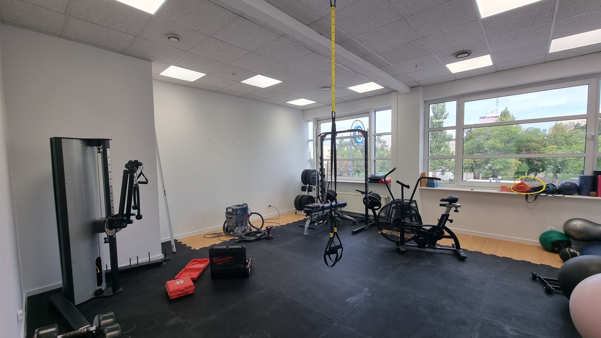 Jasne wnętrze siłowni z różnorodnym sprzętem do ćwiczeń: maszyna wyciągowa, rowerek, TRX, ciężary. Widoczne narzędzia sugerują trwające prace wykończeniowe.