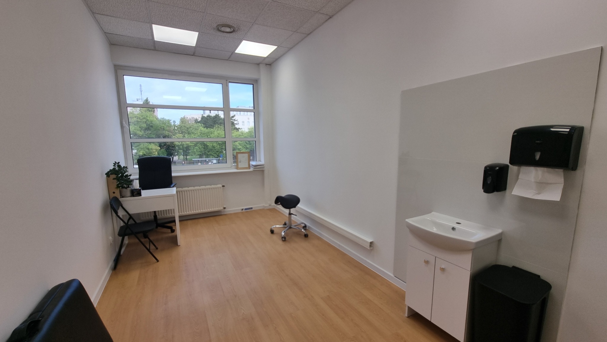 Jasne biuro z umywalką, biurkiem i krzesłem na kółkach. Widok na zieleń przez okno. Minimalistyczny design, drewniana podłoga i białe ściany.