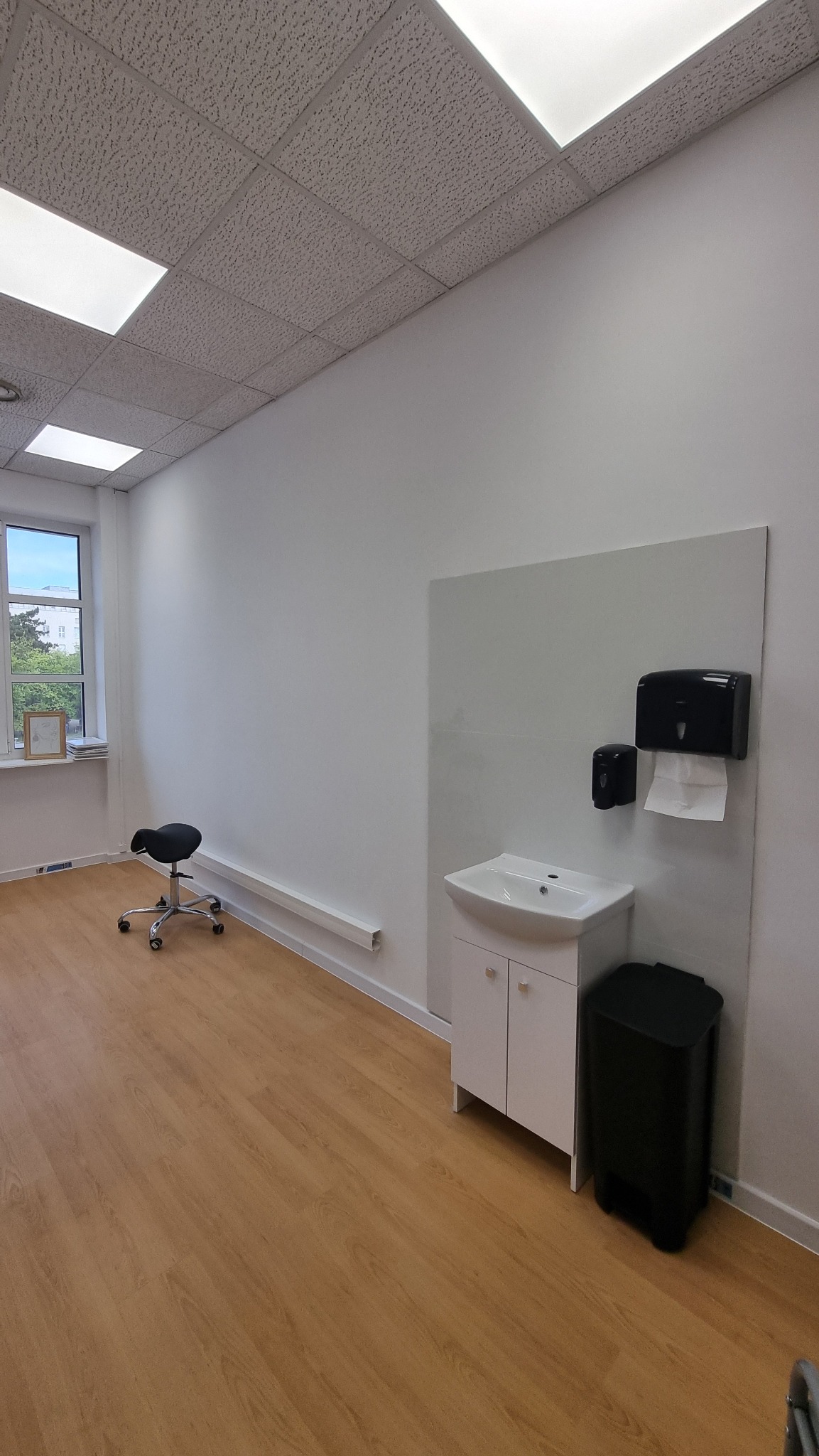 Jasne wnętrze biurowe z umywalką, krzesłem na kółkach i drewnianą podłogą. Minimalistyczny design z oknem i białymi ścianami.