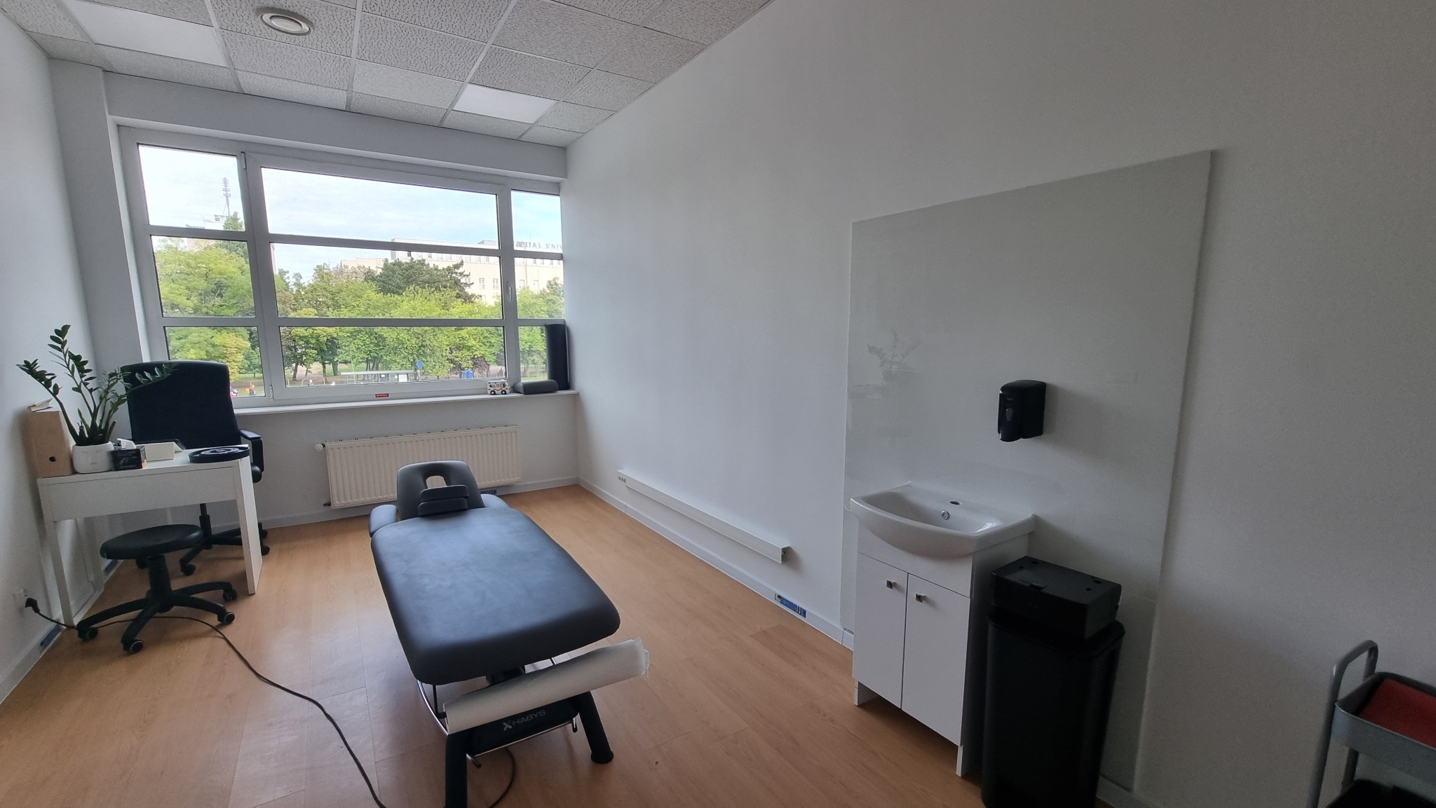 Jasne wnętrze gabinetu z kozetką do masażu, umywalką i biurkiem przy oknie. Minimalistyczny design, parkiet na podłodze, widok na zieleń za oknem.