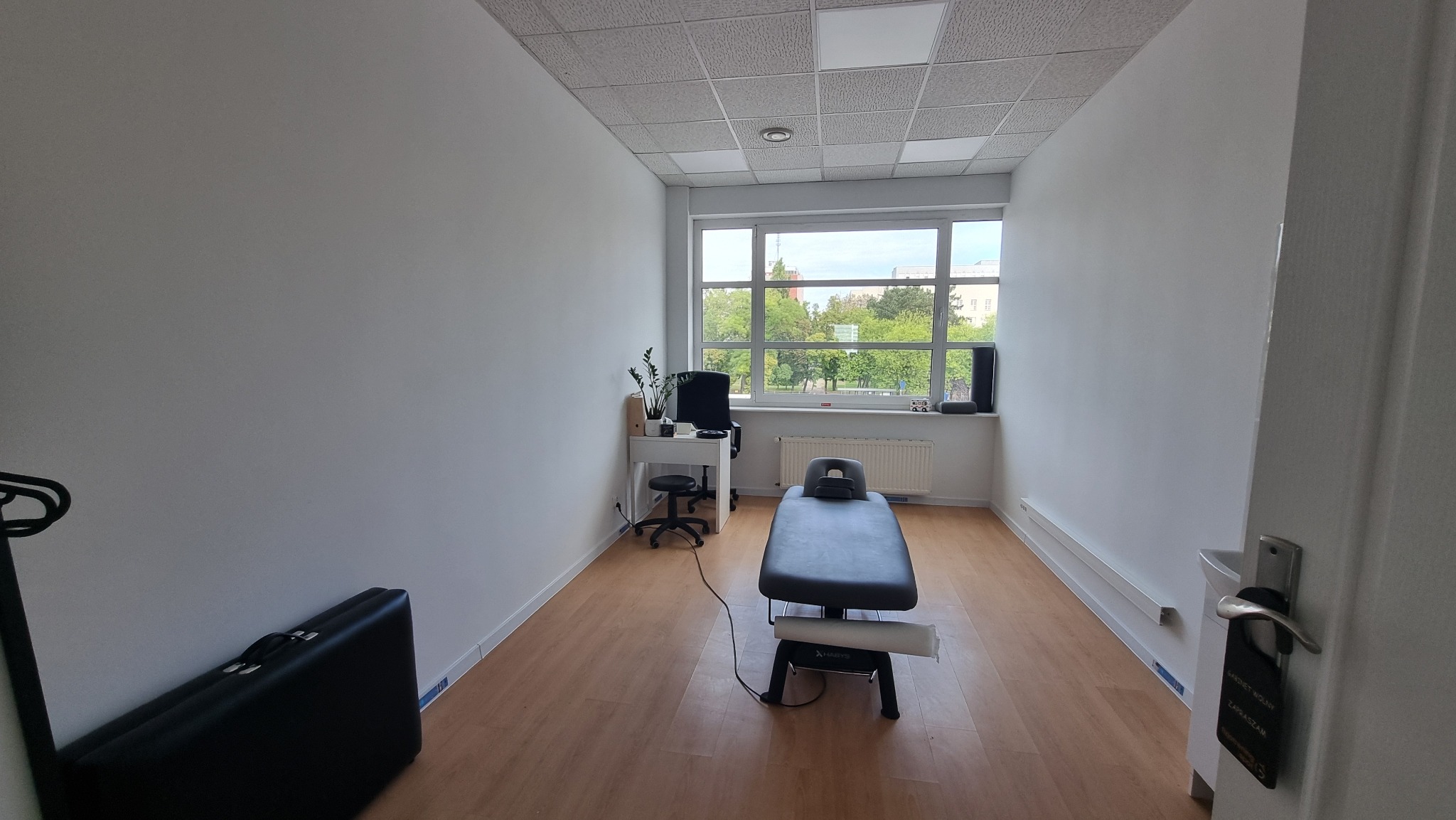 Jasne wnętrze gabinetu z kozetką do masażu, biurkiem i oknem z widokiem na zieleń. Minimalistyczny wystrój, drewniana podłoga, czyste ściany.