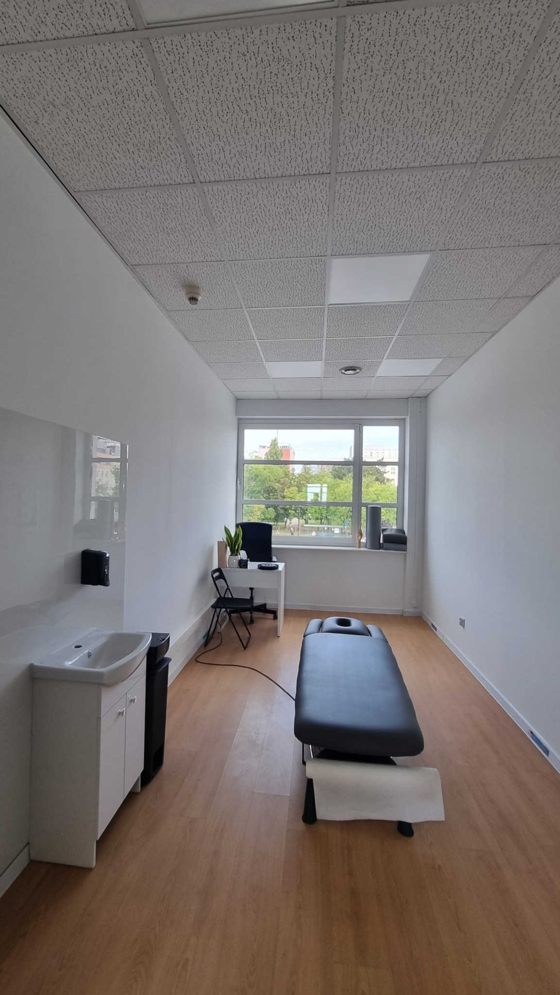 Jasne wnętrze gabinetu z kozetką, biurkiem przy oknie i umywalką. Parkiet na podłodze, sufit kasetonowy. Widok na zieleń za oknem.