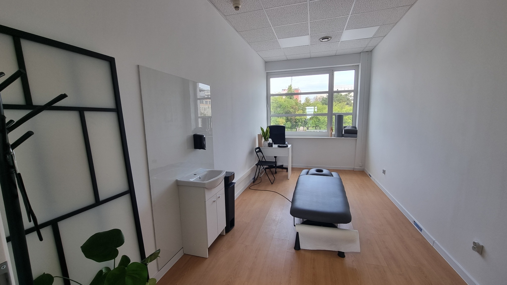 Jasne wnętrze gabinetu z kozetką do masażu, umywalką i biurkiem pod oknem. Minimalistyczny wystrój, drewniana podłoga i duża ilość światła dziennego.