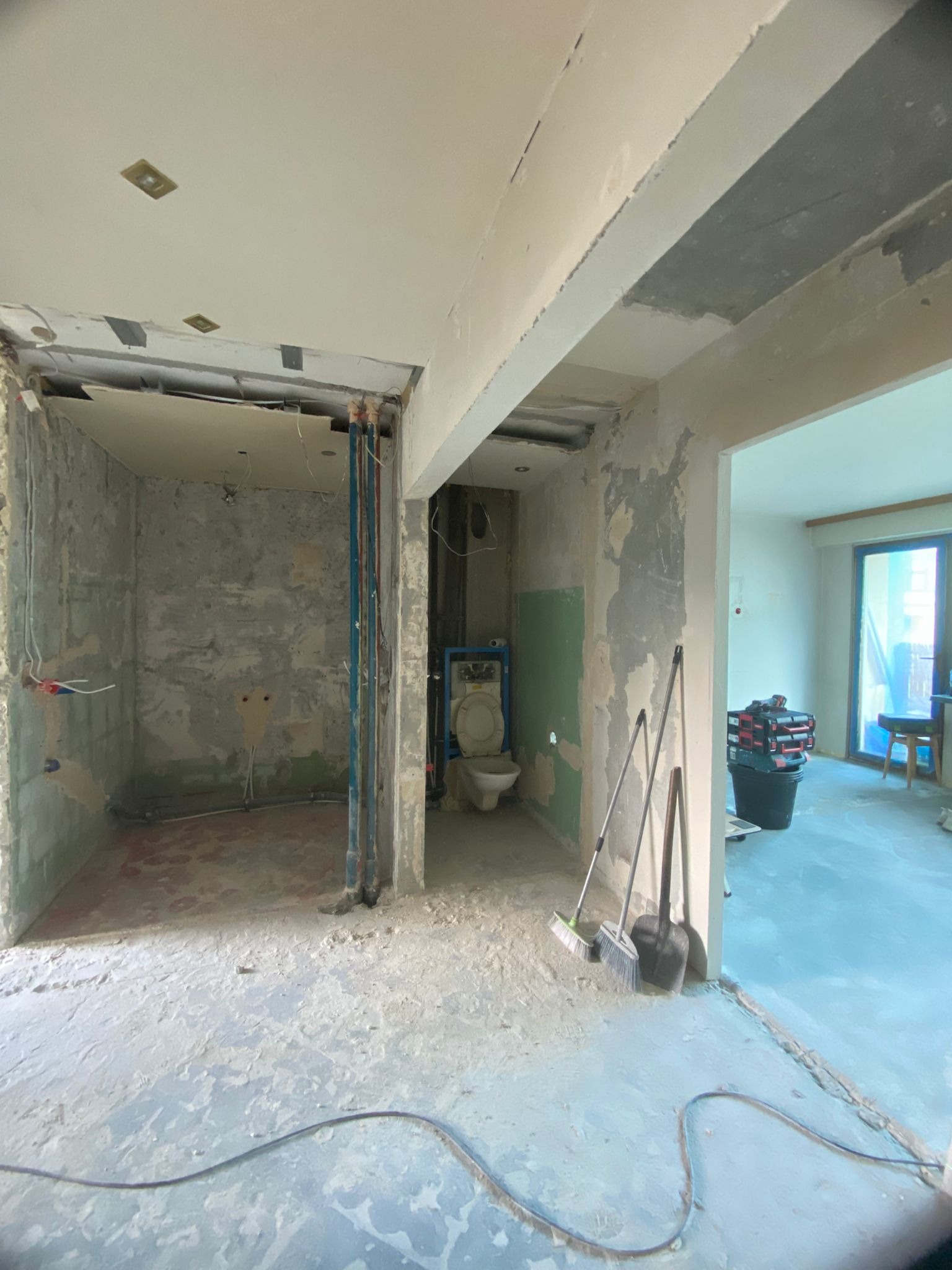 Wnętrze w trakcie prac przygotowawczych do remontu: widoczne instalacje, ściany w stanie surowym, narzędzia. W tle widoczny pokój z oknem.