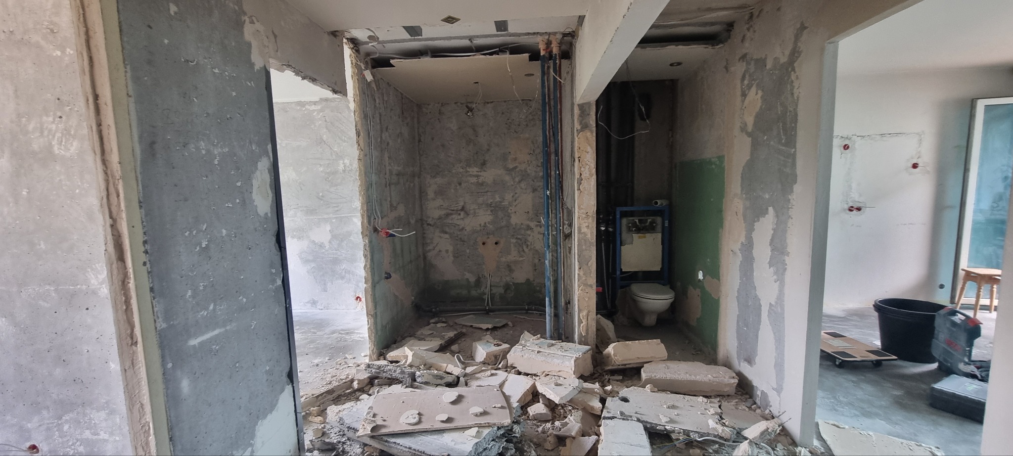 Łazienka w trakcie remontu: surowe ściany, gruz na podłodze, widoczne instalacje i stelaż WC. W tle wiadro i narzędzia.