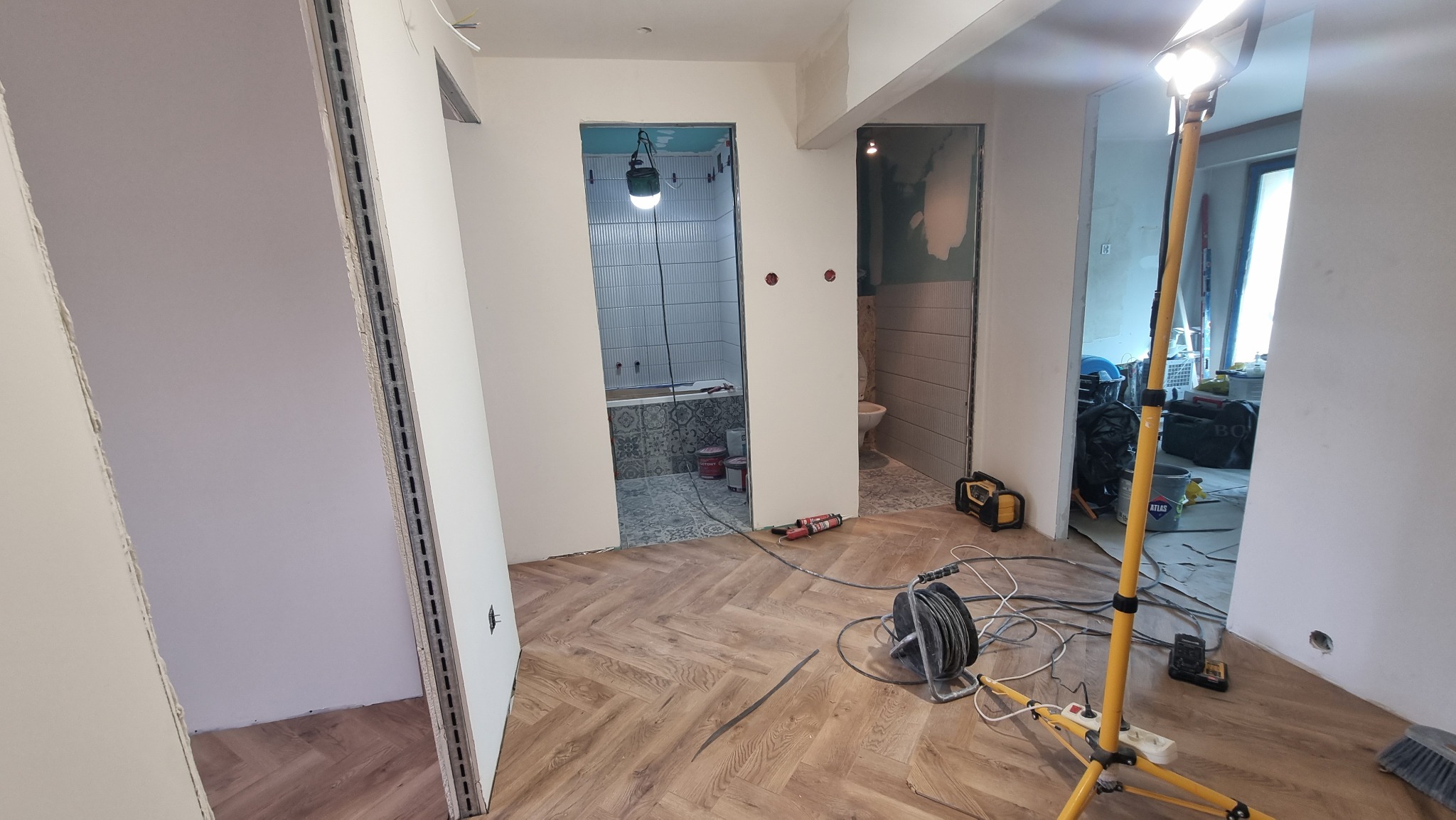 Wnętrze w trakcie prac wykończeniowych: widok na łazienkę z ułożonymi płytkami, ułożona podłoga, ściany przygotowane do malowania, narzędzia pracy i oświetlenie.