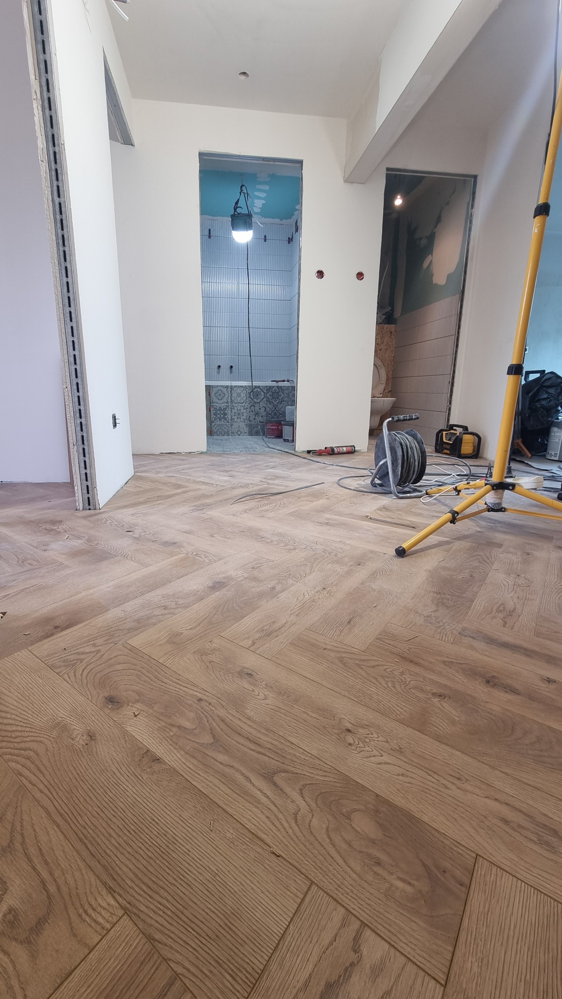 Parkiet ułożony w jodełkę na podłodze w trakcie remontu. W tle widoczna łazienka w trakcie wykończenia, z położonymi płytkami. Na podłodze leżą narzędzia.