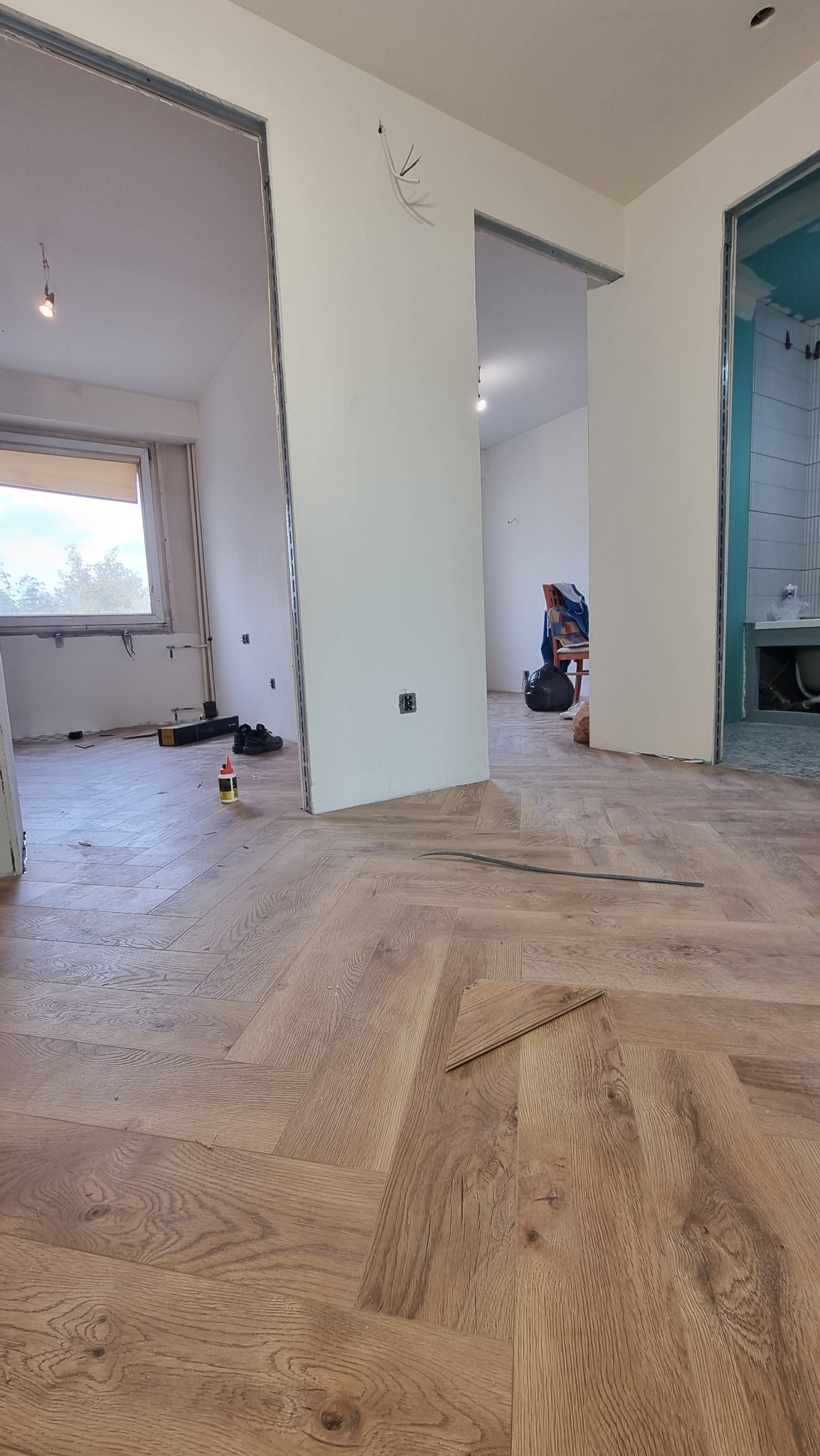 Ułożona podłoga z paneli w jodełkę w remontowanym mieszkaniu. Widoczne narzędzia, klej i fragment niezakończonej podłogi. W tle wykończona łazienka.