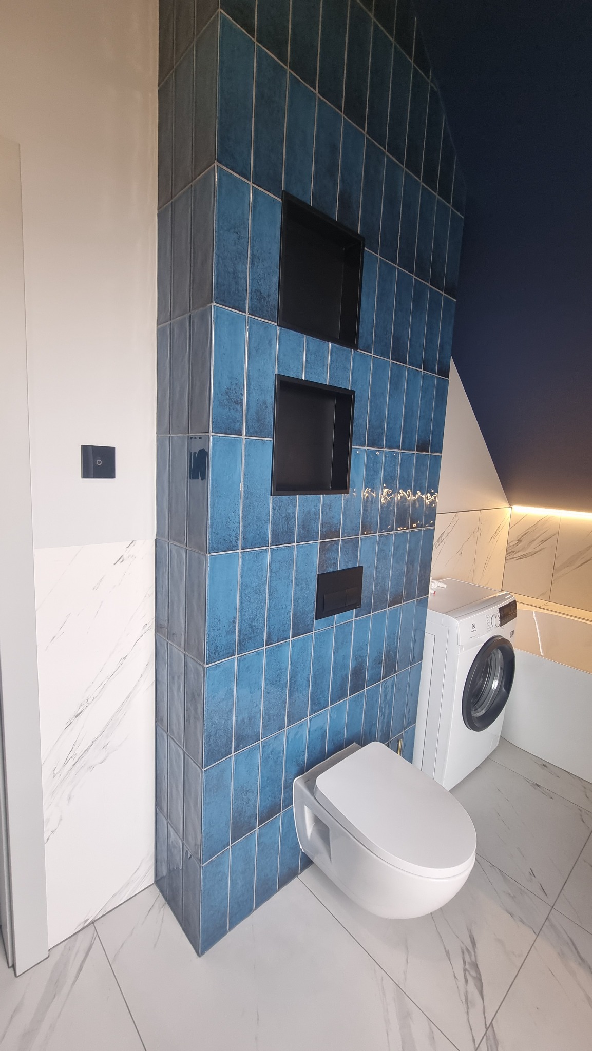Niebieska, pionowa mozaika w łazience z wnękami, wisząca toaleta i pralka. Marmurowe płytki podłogowe i ściany. Nowoczesny design z akcentem kolorystycznym.