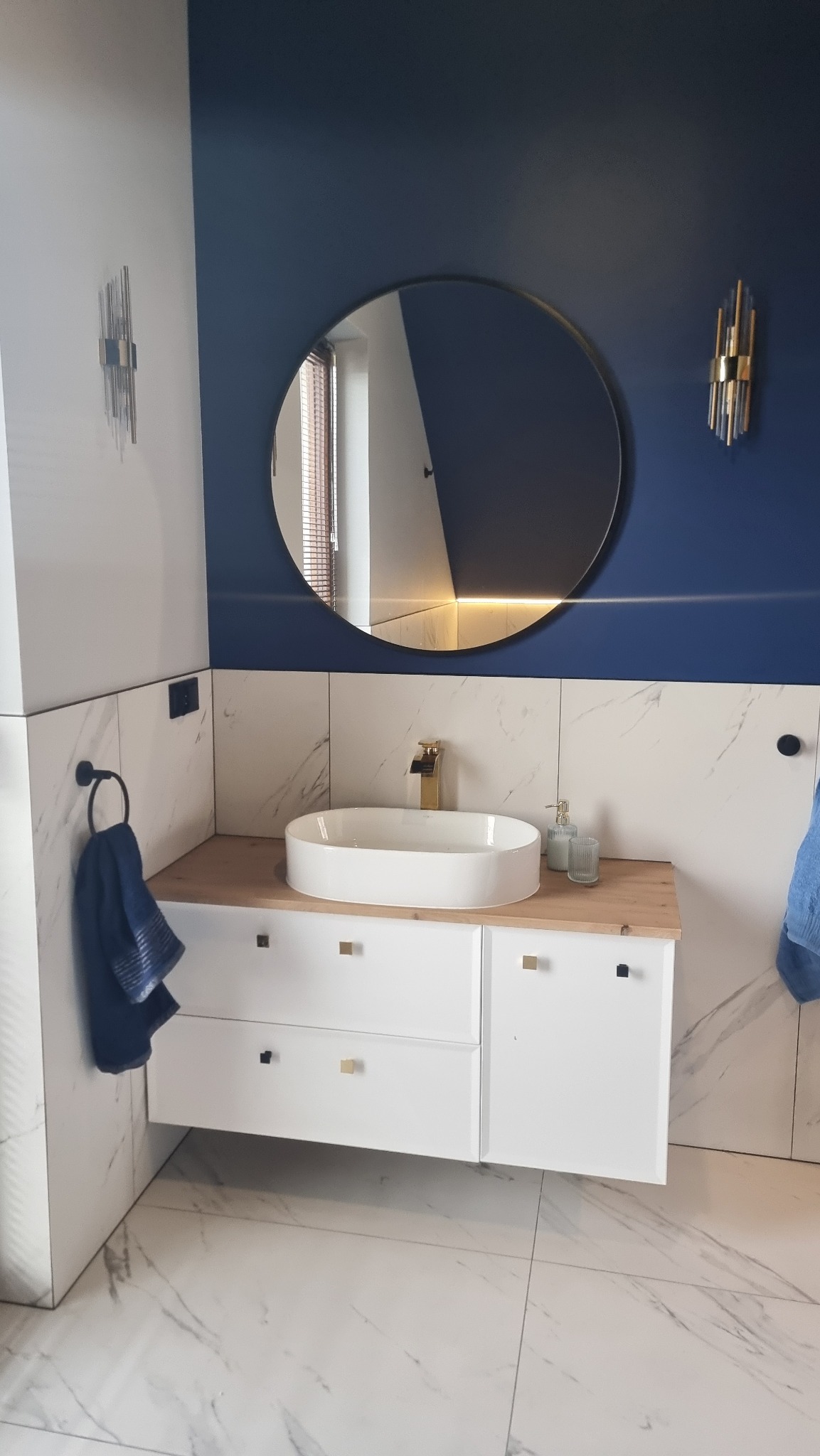 Nowoczesna łazienka z owalną umywalką nablatową, złotą armaturą i okrągłym lustrem na tle granatowej ściany. Białe płytki imitujące marmur i biała szafka pod umywalką.